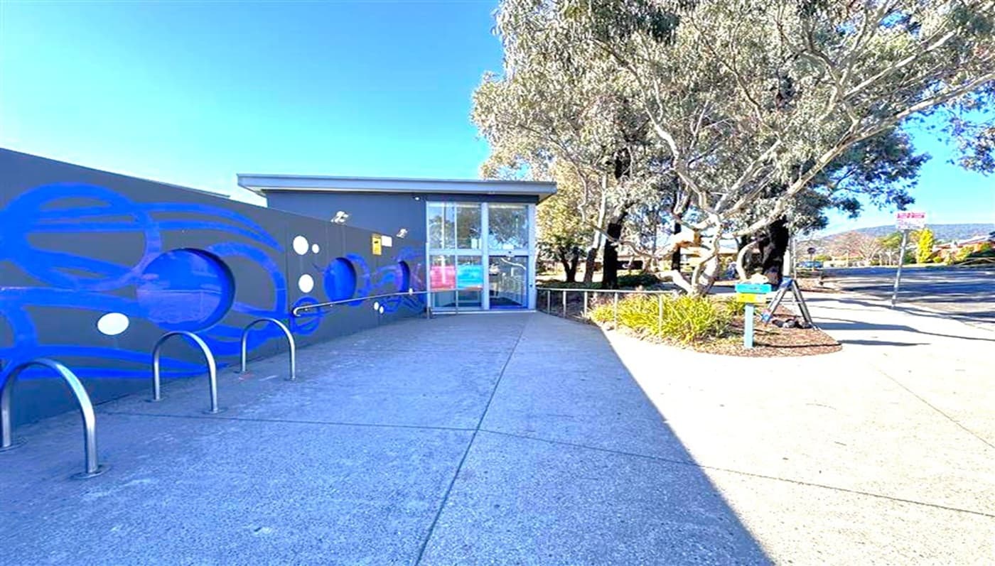 A building with a modern design is on the left, featuring a dark wall with circular blue and white patterns. In front of the wall, there are metal bike racks. The entrance has glass doors. On the right, there are trees with a small landscaped area and a signpost. The ground is paved with concrete, and the sky is clear.