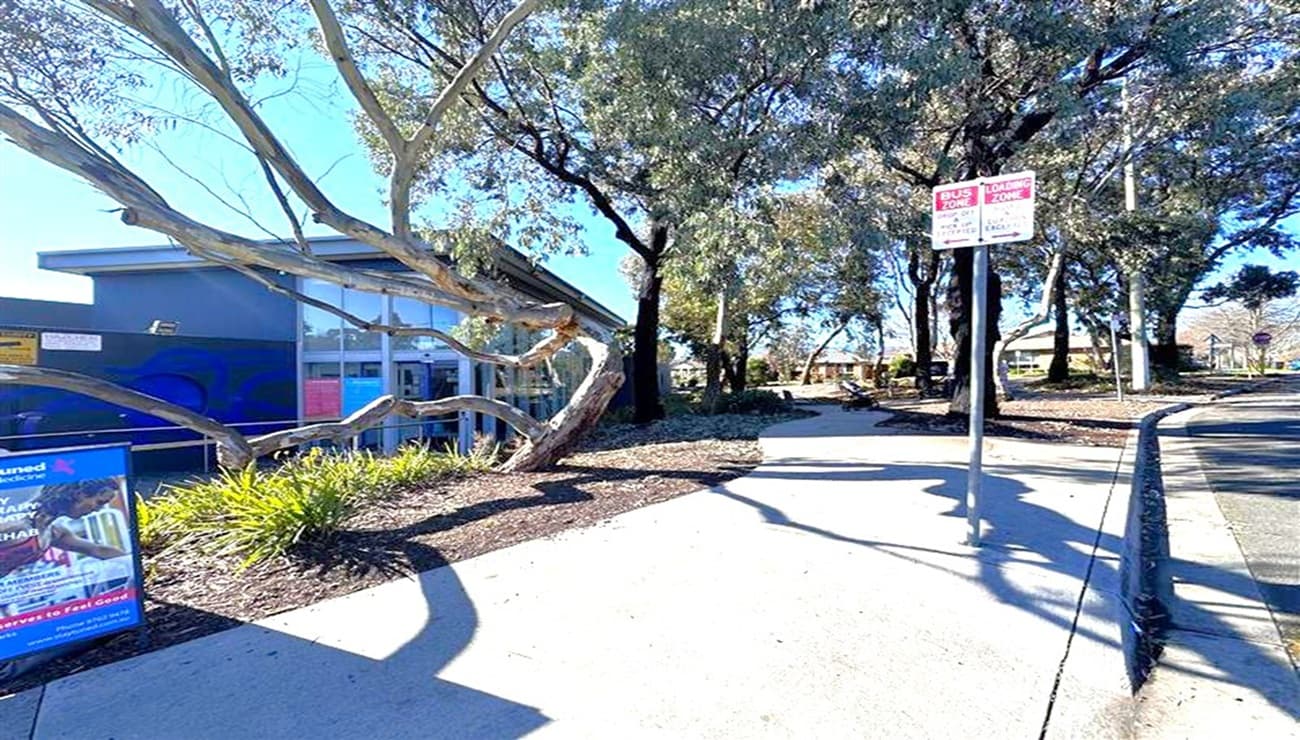 A footpath runs through the centre, bordered by a garden bed with shrubs and a large, twisting tree on the left. A blue building with large windows is partially visible behind the tree. On the right, a signpost with red and white signage stands next to the path. In the background, more trees and a glimpse of a building can be seen. The ground is paved, and the area is well-shaded by the trees.