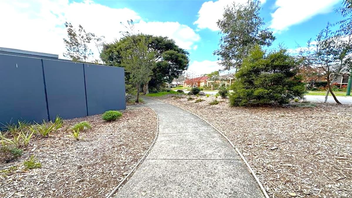 A narrow concrete path curves through a landscaped area. On the left, there's a dark blue wall with small plants and mulch at its base. On the right, various trees and shrubs are scattered across a mulched area. In the background, a row of houses is visible, partially obscured by trees. The sky is partly cloudy.