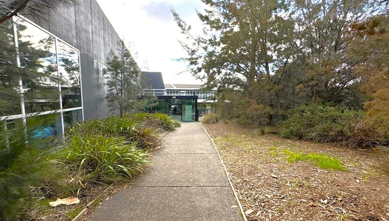 A narrow path runs through the centre, bordered by green grass and mulch. On the left, a building with large glass windows reflects the surroundings. On the right, dense foliage and trees create a natural barrier. In the background, a structure with a metal roof and a glass entrance is visible. The sky is partly cloudy.
