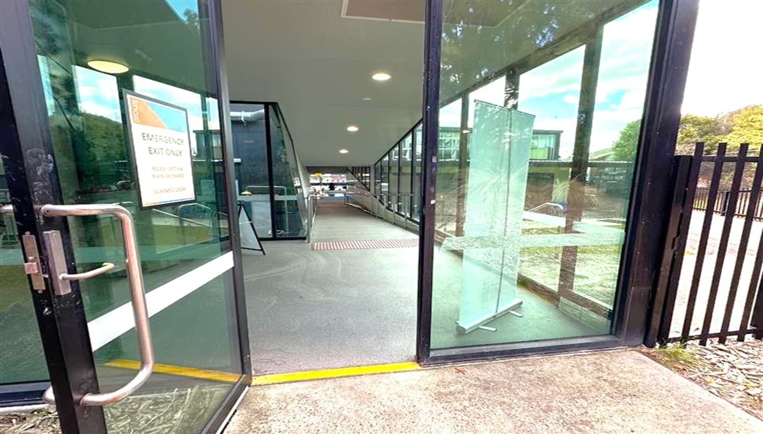 Glass doors open to a corridor with a grey floor and a ceiling with recessed lights. On the left, a sign on the door reads "Emergency Exit Only." The corridor is lined with large windows on the right, offering a view of a fenced outdoor area with trees and a parked car. A transparent stand is positioned near the entrance. The corridor extends into the distance, leading to another area inside the building.