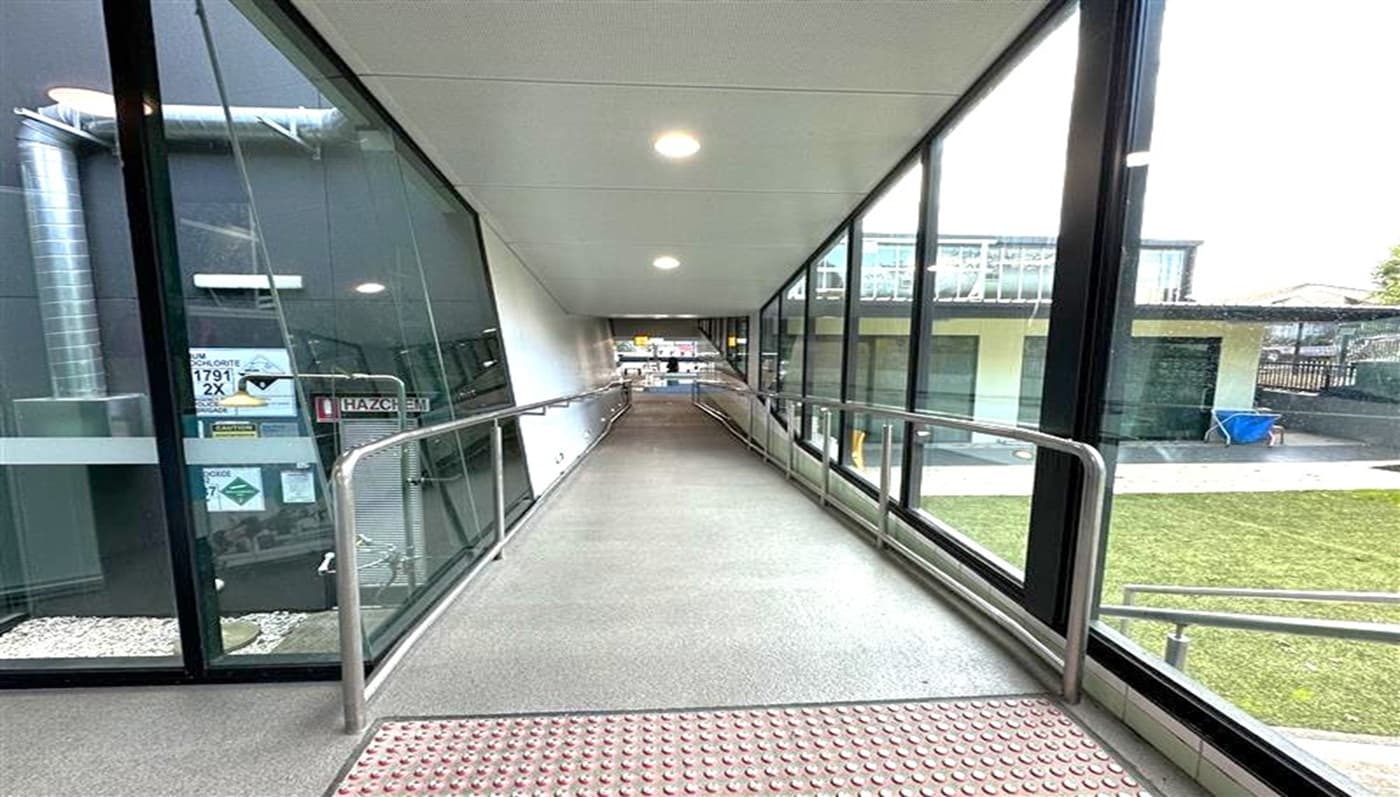 A covered walkway with a gentle ramp, featuring metal railings on both sides. The left side has large glass windows showing an industrial area with pipes and signs. The right side also has glass windows, revealing a grassy outdoor area and a building with green doors. The ceiling is white with recessed lights, and the floor is grey with a textured section at the start of the ramp.