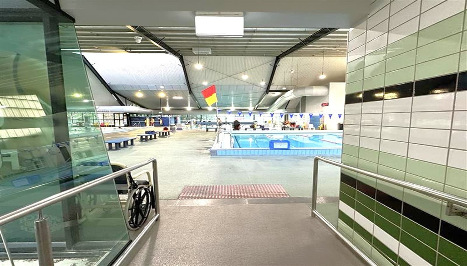 Entrance to an indoor swimming pool area. On the left, there's a glass wall and a metal railing. The right side features tiled walls with black horizontal stripes. In the centre, a pool with a red and yellow flag is visible, surrounded by a deck with benches. The ceiling is high with exposed beams and lights. The floor is grey and textured.