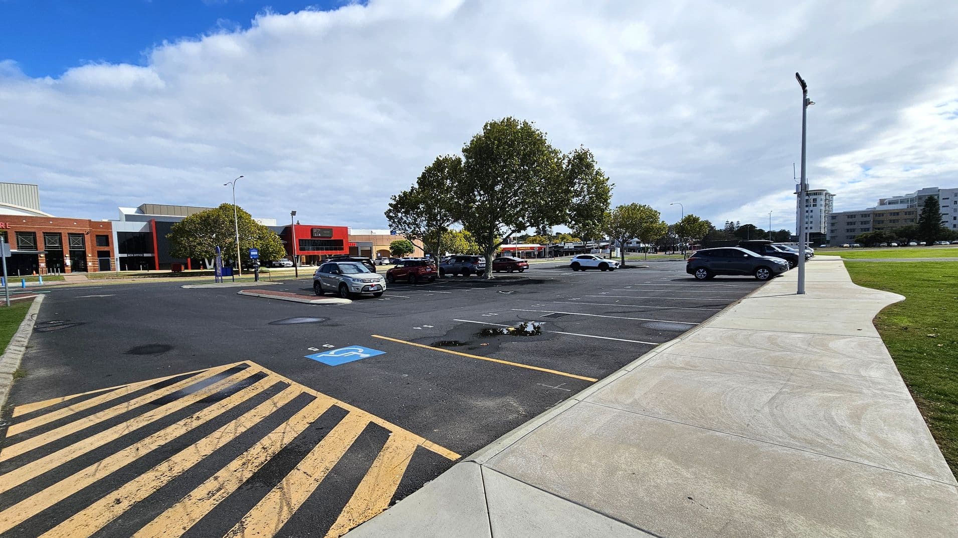 A car park with several vehicles is visible, with a row of trees in the centre. On the left, there is a brick building with a red and black facade. The foreground shows a pedestrian crossing with yellow and black stripes and a disabled parking space. On the right, a concrete footpath runs alongside the parking spaces, leading to a grassy area. In the background, there are more buildings, including a tall white one. A streetlamp is positioned near the footpath.