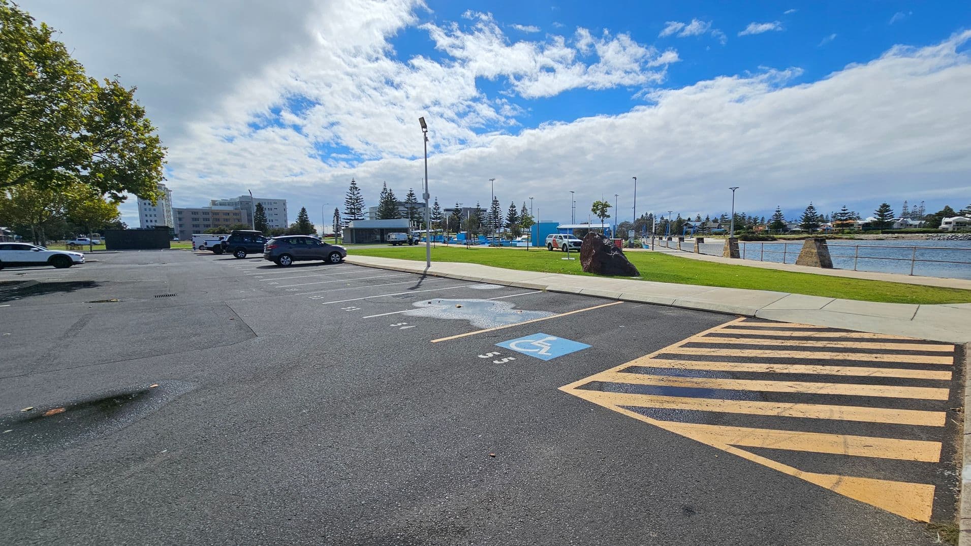 A car park with marked spaces, including a designated disabled parking spot with a blue symbol, is in the foreground. To the right, a yellow-striped area is visible. In the background, there is a grassy area with a large rock, a few trees, and a body of water with a railing along the edge. Several buildings and parked cars are on the left, with a tree providing some shade. The sky is partly cloudy.