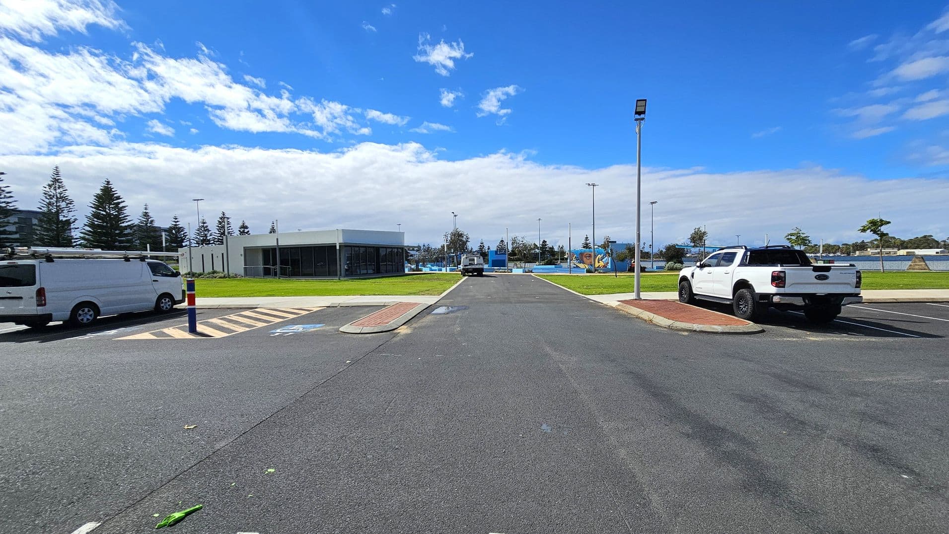 A car park with a white van on the left and a white pickup truck on the right. In the centre, a road leads to a building with large windows and a flat roof. The background features a grassy area with scattered trees and a playground. A tall streetlight is on the right side of the road. The sky is partly cloudy.