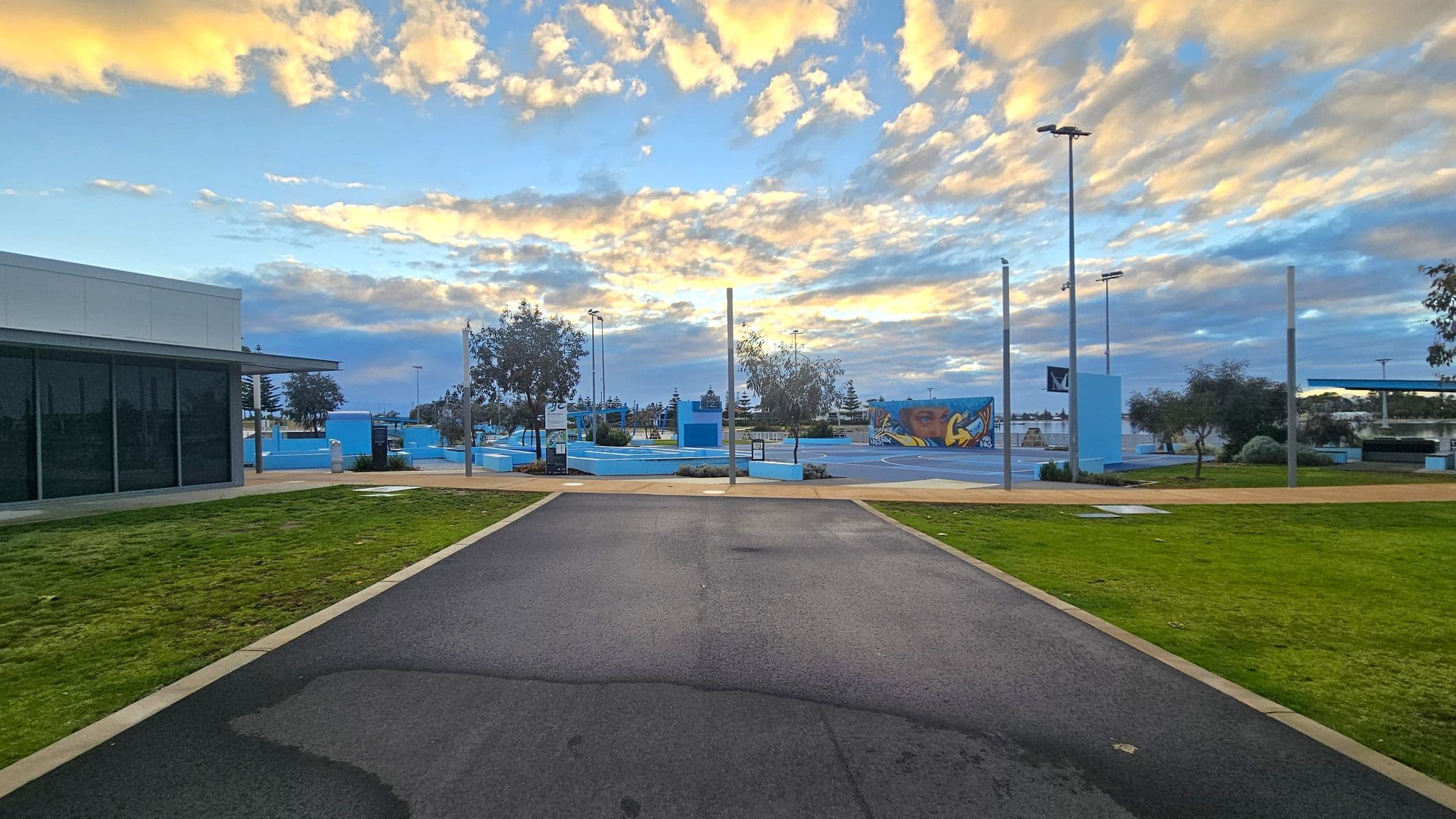 A paved path leads towards a skate park in the centre, with blue ramps and a colourful mural. On the left, a building with large dark windows and a flat roof. On the right, green grass and scattered trees. Tall light poles are visible throughout the area. The sky is filled with clouds tinged with yellow and orange hues.