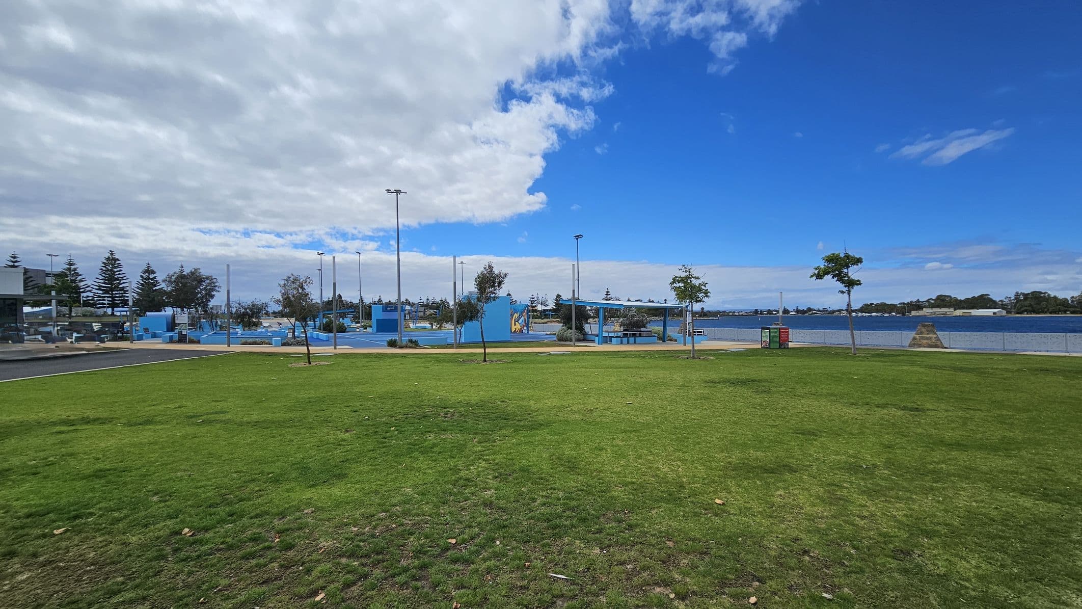A grassy park area with patches of green and brown in the foreground. In the middle, there are several small trees and a pathway leading to a blue-painted recreational area with structures and poles. To the right, a body of water is visible, bordered by a fence. The background features more trees and a partly cloudy sky.