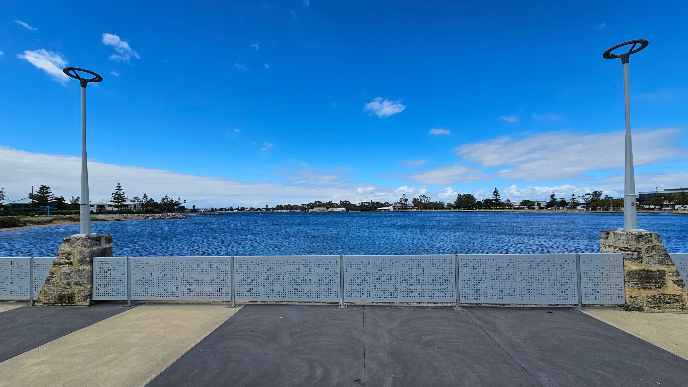 A waterfront scene with a wide expanse of blue water in the middle. In the foreground, there is a paved area with alternating light and dark sections. Two modern street lamps with circular tops are positioned on stone bases, one on the left and one on the right. A metal railing with a perforated design runs across the middle. In the background, there are trees and buildings along the shoreline under a clear blue sky.