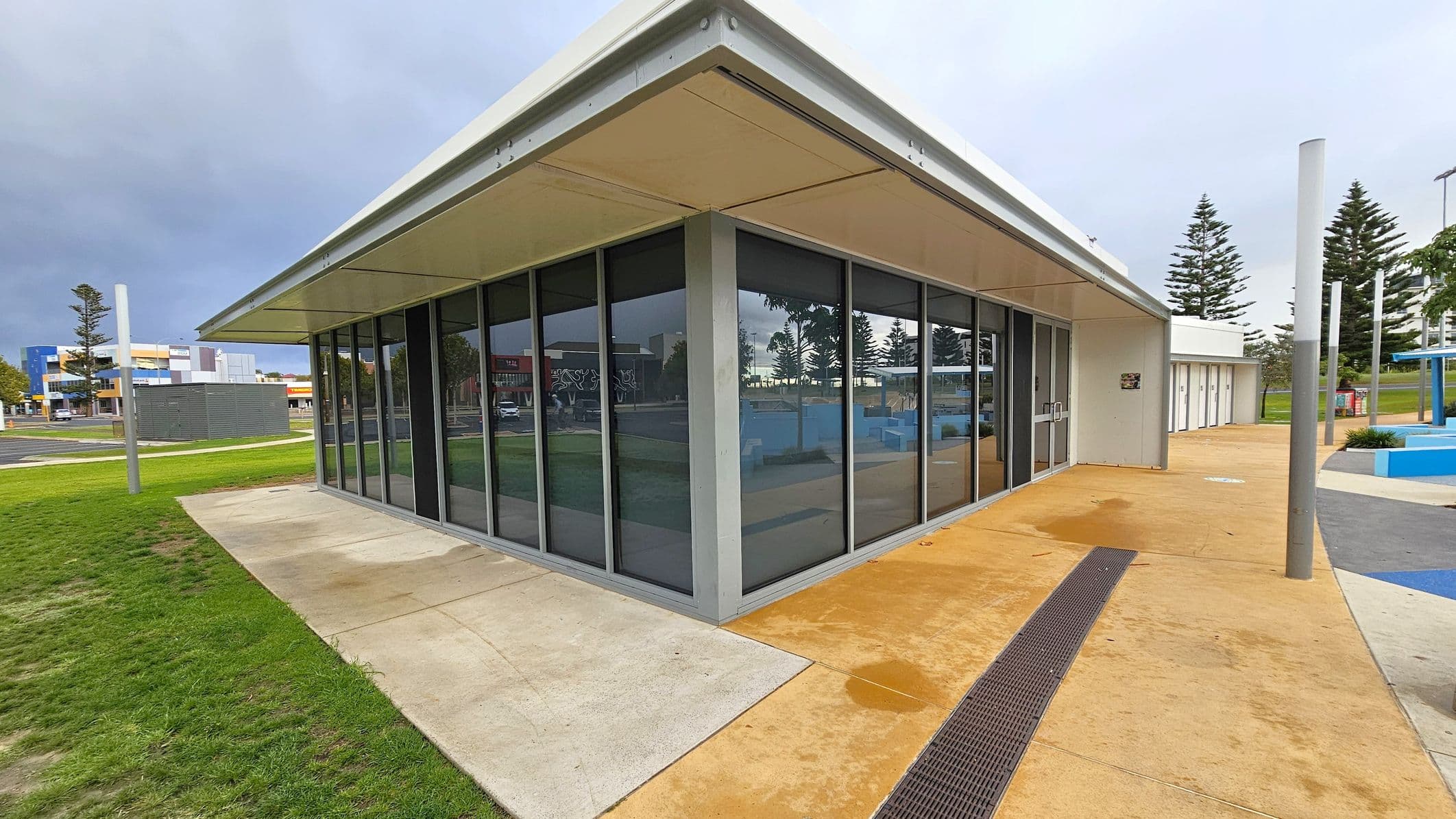 A modern building with large glass windows on the left, set on a concrete path that transitions into grass. The right side features a continuation of the building with a beige walkway and a series of doors. Tall, slender poles are positioned along the path, and several pine trees are visible in the background. The ceiling is flat and white, matching the building's exterior.