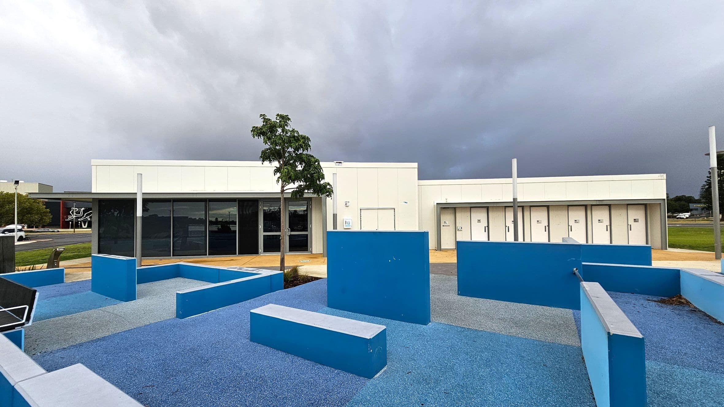 A modern building with a flat roof and white exterior is in the background. In front, there is a playground area with blue rubber flooring and various blue geometric structures, including benches and walls. A small tree is planted near the centre. To the left, a road with a few cars and a building with a mural can be seen. The sky is overcast, creating a grey backdrop.