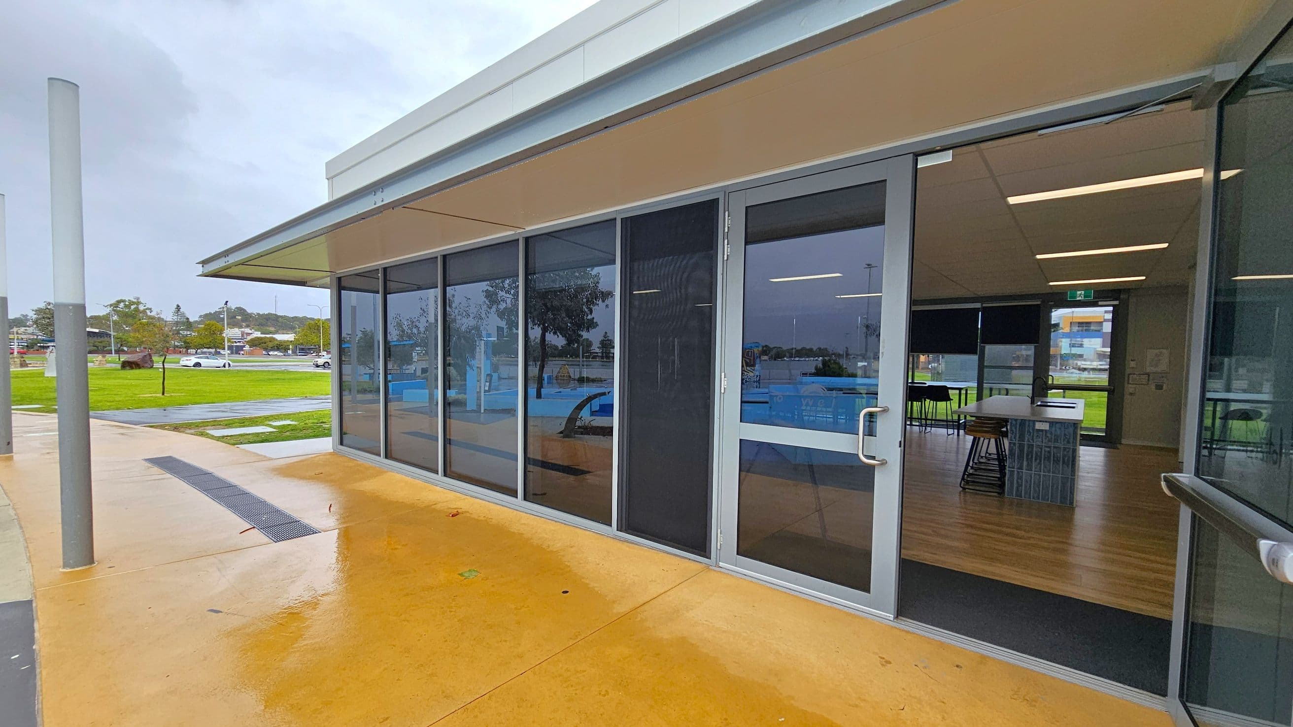A modern building with large glass windows and doors on the right, reflecting the surroundings. Inside, there are tables and chairs on a wooden floor. The ceiling has recessed lighting. Outside, the ground is wet, suggesting recent rain, with a yellowish concrete surface. In the background, there is a grassy area with trees and parked cars. A tall, cylindrical post is visible on the left.