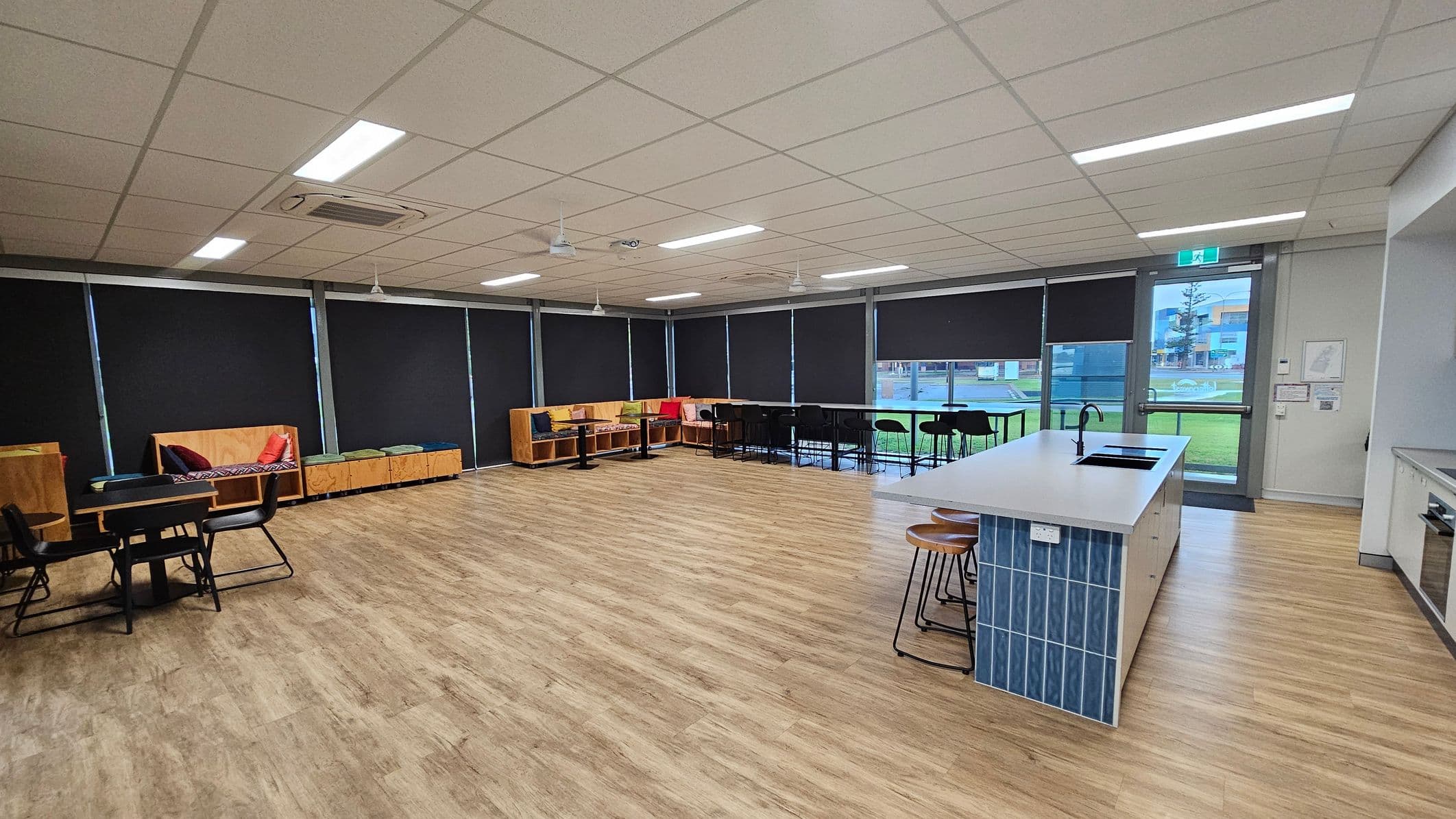 A spacious room with a light wooden floor and a white tiled ceiling. On the left, there are tables with black chairs and wooden benches with colourful cushions. The right side features a kitchen island with blue tiles and wooden stools. Large windows with black blinds line the back wall, and a glass door leads outside. The room is well-lit with fluorescent lights.