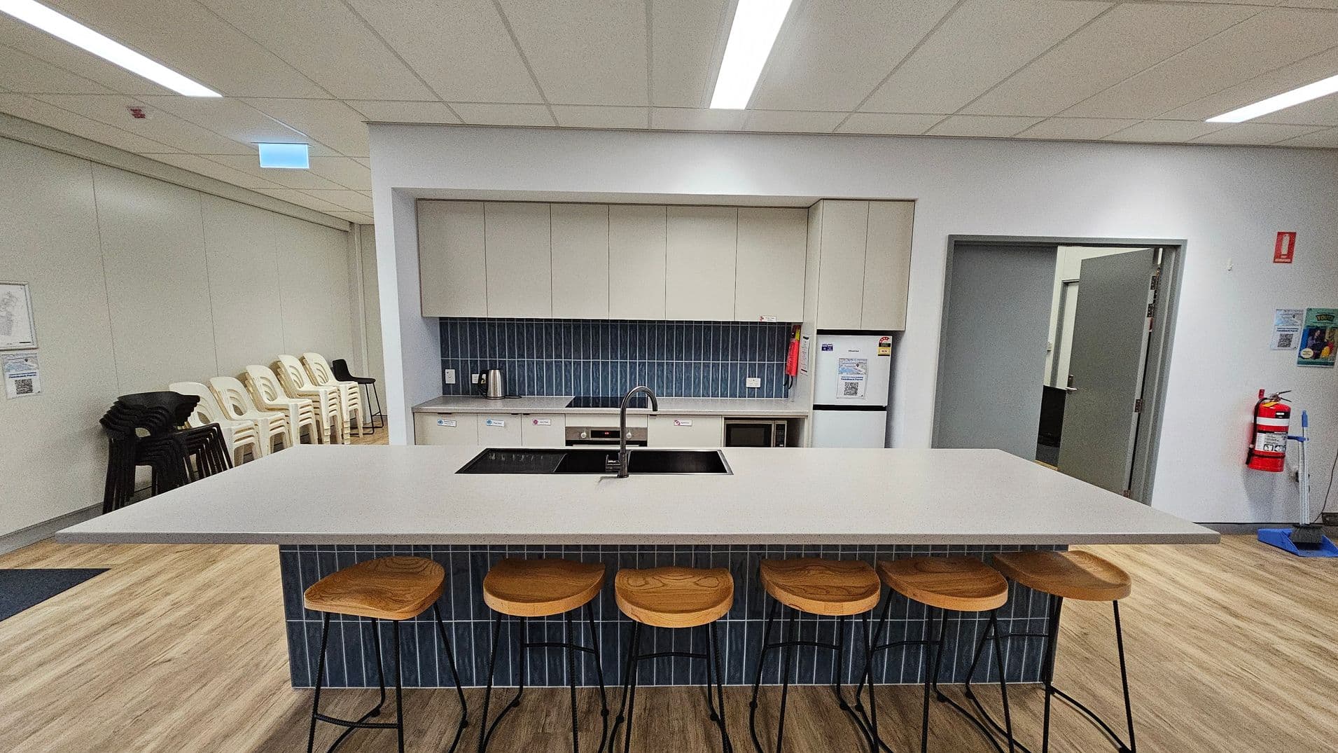 A modern kitchen area with a large island in the centre featuring a white countertop and a built-in sink. Five wooden bar stools with metal legs are lined up in front. The back wall has light-coloured cabinets and a blue-tiled backsplash. Appliances include a kettle, stovetop, and a fridge. To the left, stacked white and black chairs are against the wall. On the right, a fire extinguisher is mounted near an open door. The floor is wooden, and the ceiling has recessed lighting.
