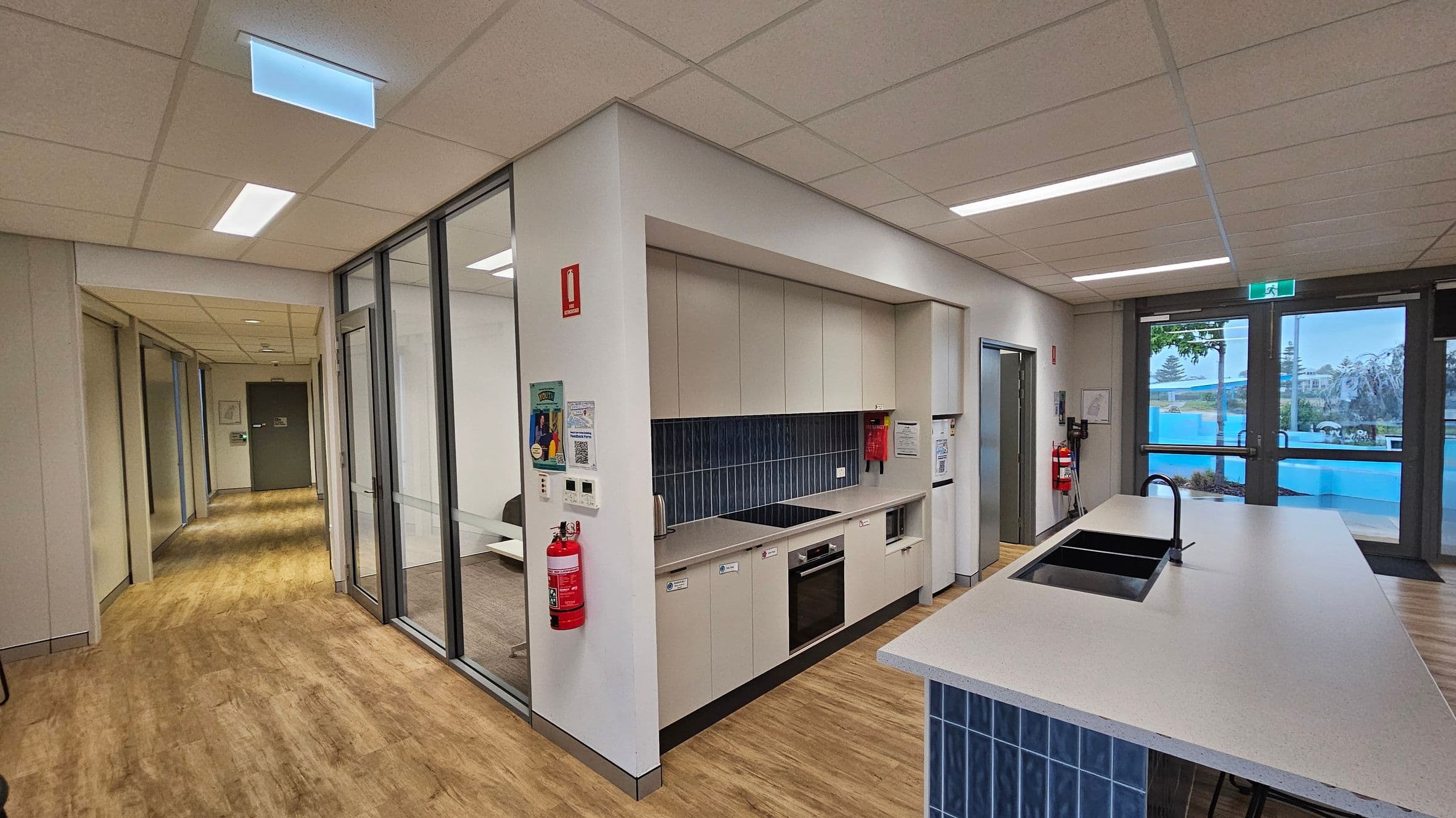 A modern kitchen area with light wood flooring and a suspended ceiling with recessed lighting. On the left, there is a hallway leading to a green door. In the centre, a kitchenette with beige cabinets, a black cooktop, and a built-in oven. A fire extinguisher is mounted on the wall. On the right, a large island with a sink and tap, and glass doors leading outside. Blue tiles are used as a backsplash and on the island base.