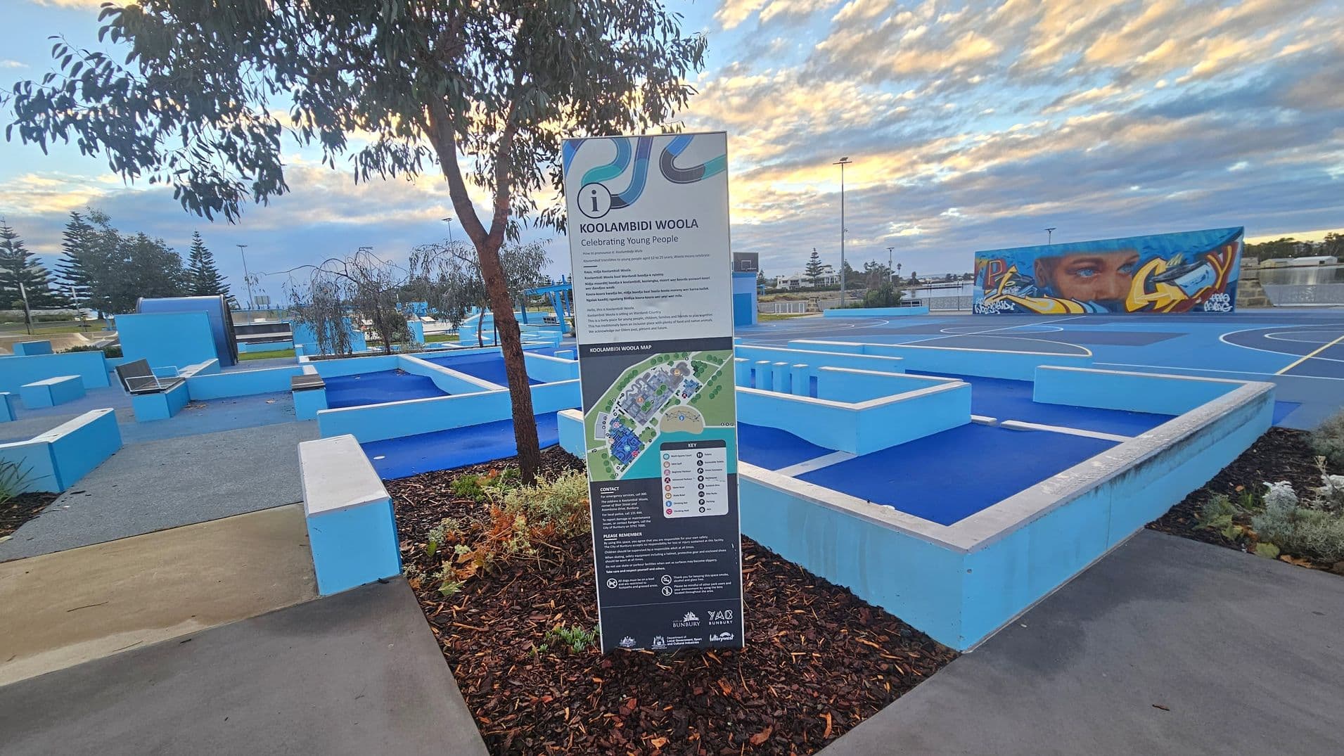 A park area with a prominent sign in the centre detailing "Koolambidi Woola" and a map. The ground is covered with blue and grey pathways and seating areas. On the right, there is a blue sports court with a mural on a wall depicting a face and abstract designs. Trees and plants are scattered throughout, and the sky is filled with clouds.