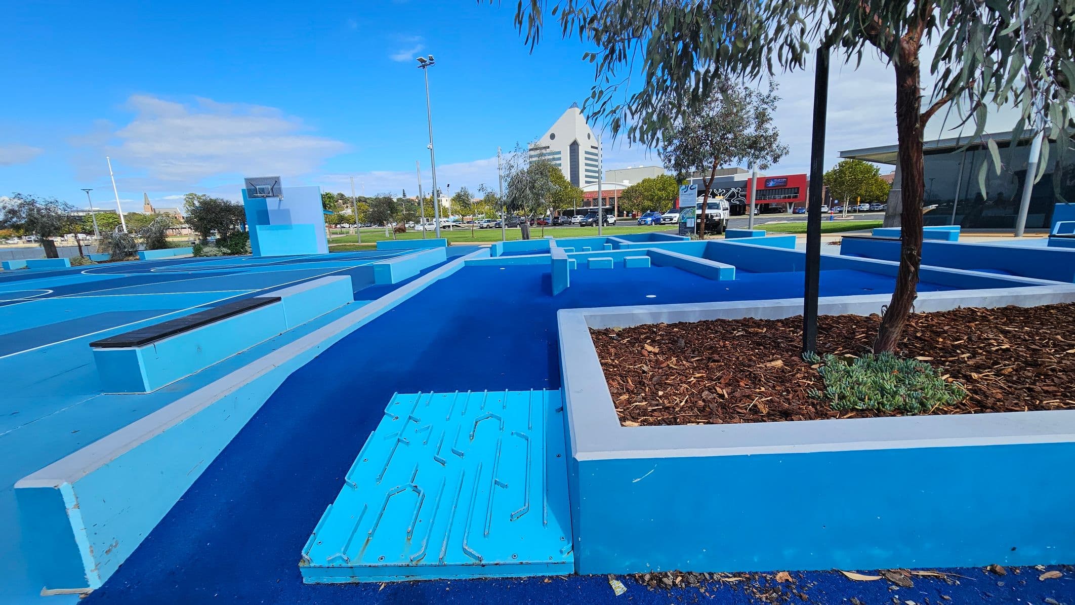 A vibrant blue outdoor area with geometric structures and pathways. On the right, a tree is planted in a raised bed filled with mulch. In the background, a distinctive triangular-roofed building is visible, along with other buildings and parked cars. The ground is covered in a textured blue surface, and there are several light poles scattered throughout the area.