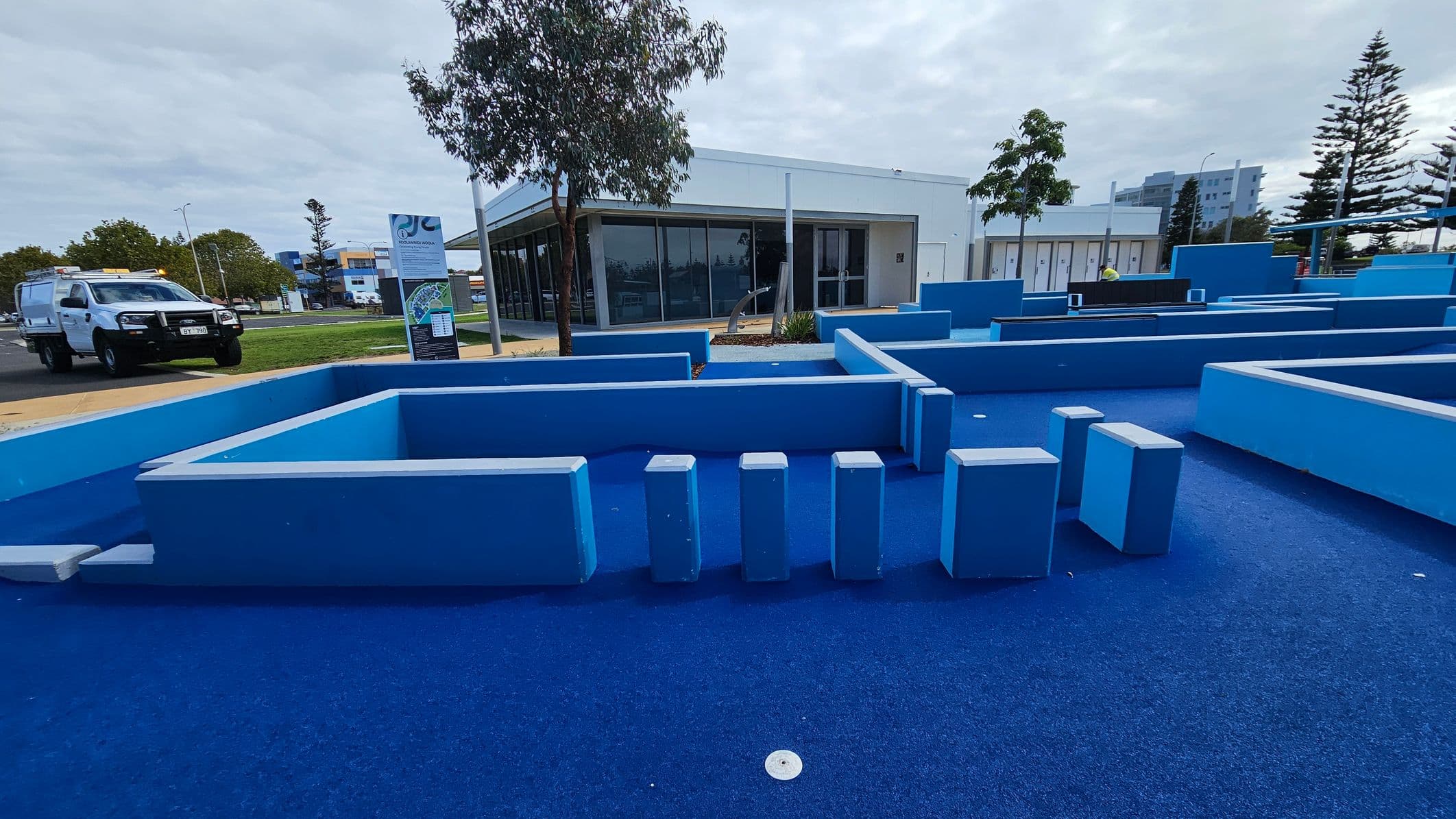 In the foreground, there is a blue outdoor play area with various geometric structures and stepping blocks. The ground is covered in blue material. In the background, a white building with large windows is visible, surrounded by a few trees. To the left, a white utility vehicle is parked on a paved area. There are signs and a grassy area near the building. Tall trees and a multi-storey building can be seen in the distance.