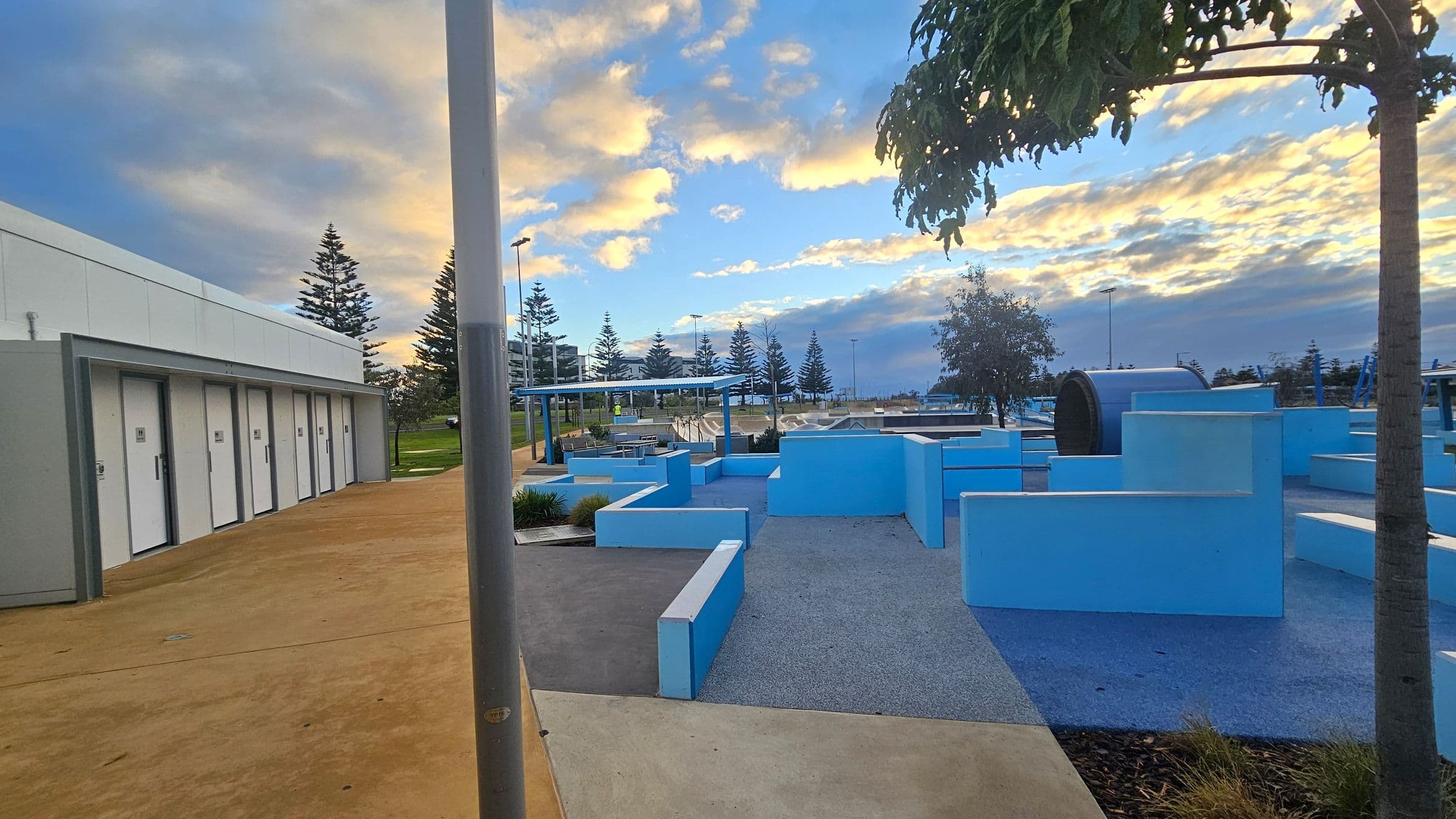 On the left, there is a row of white doors set into a grey building with a light brown concrete path in front. In the centre, a series of light blue geometric structures form a playground or park area, with a tree providing some shade. On the right, more blue structures and a large cylindrical object are visible. The background features several tall pine trees and a covered seating area. The sky is filled with clouds tinged with orange and blue hues.