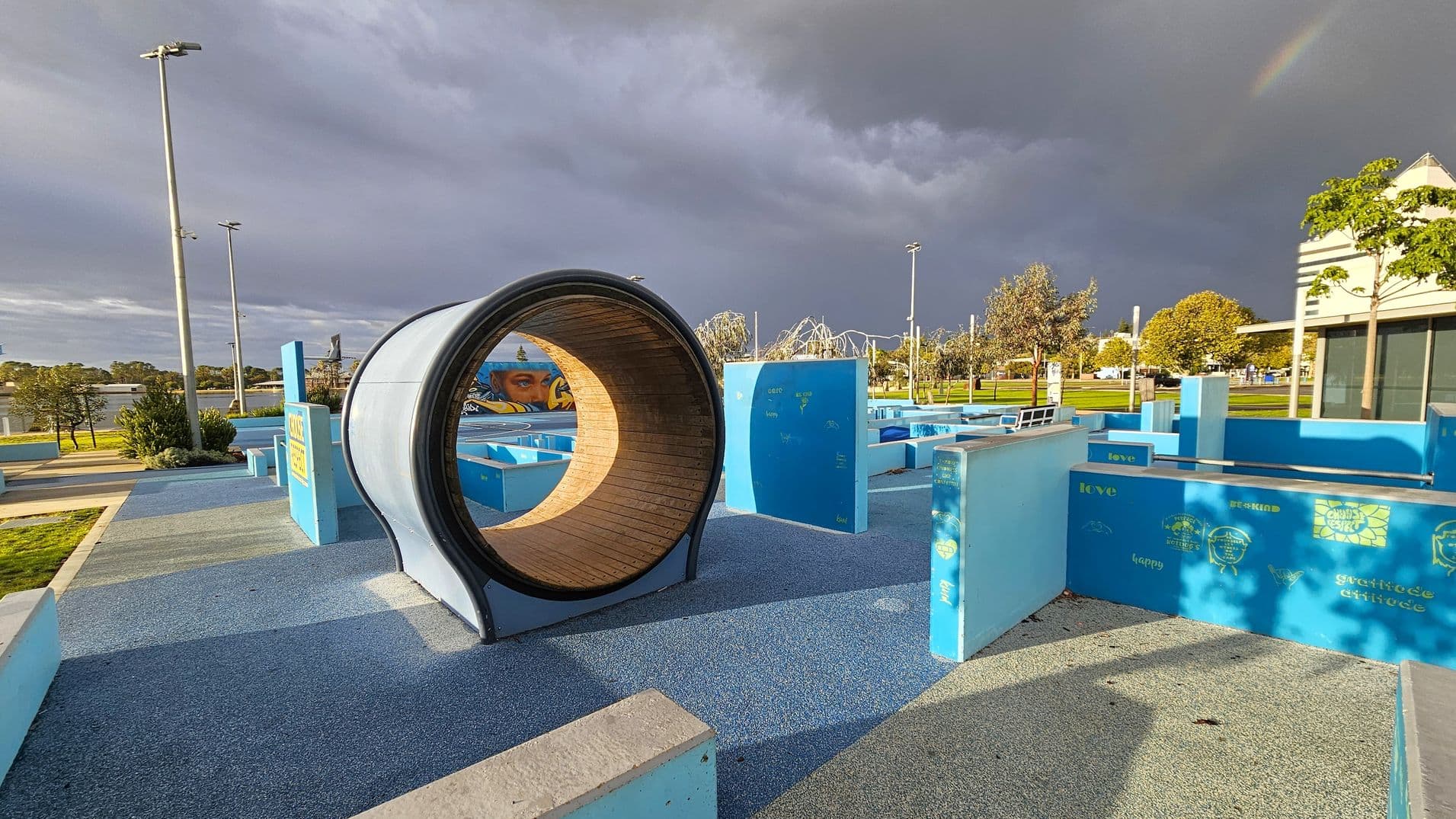 A playground with a large cylindrical tunnel in the centre, made of wood and metal. The ground is covered with blue and grey rubberised flooring. On the right, there are blue structures with words like "love" and "happy" printed on them. In the background, there are trees, a mural, and a building with a sloped roof. Tall street lamps are visible on the left, and the sky is overcast with a faint rainbow.
