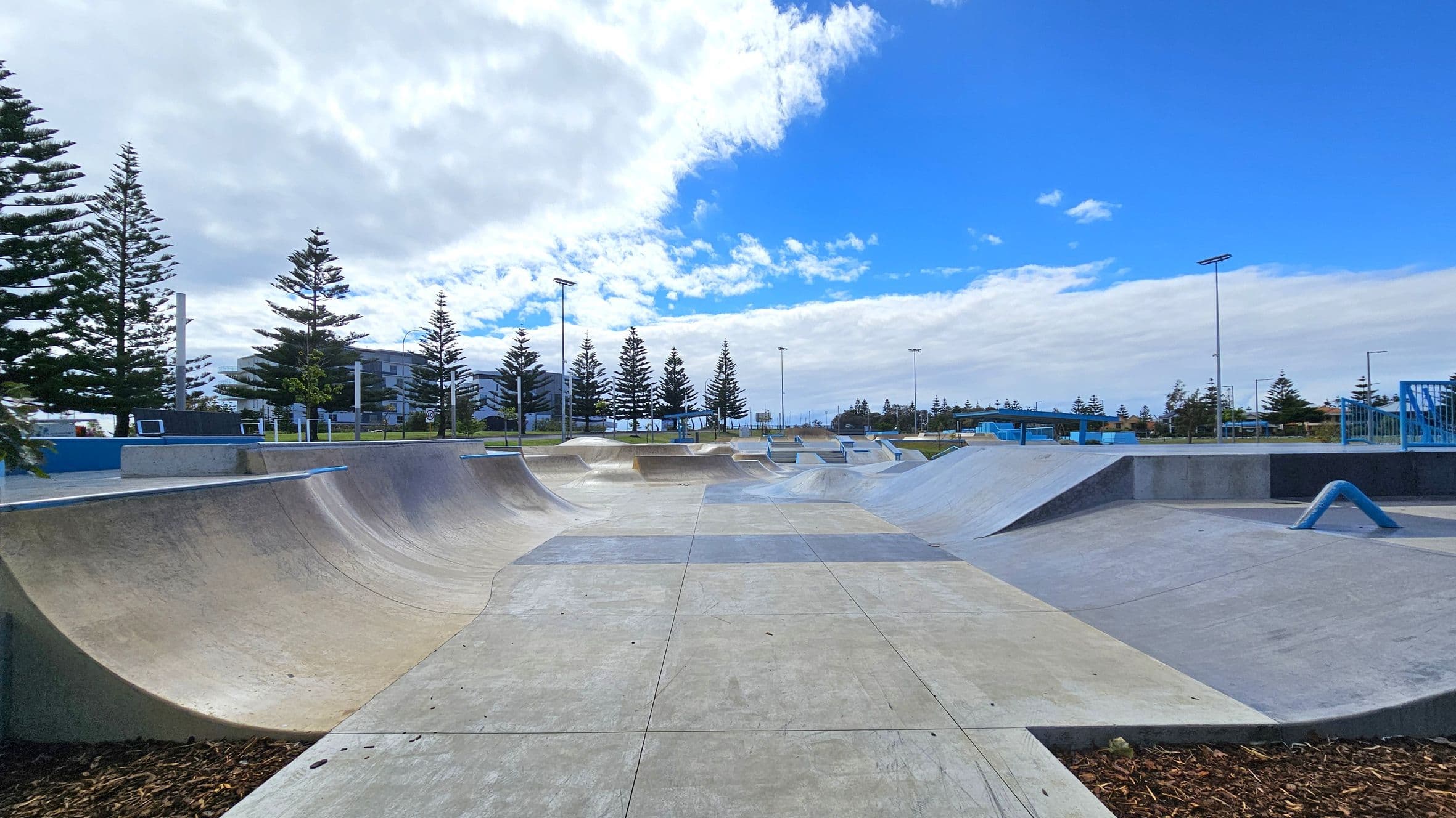 A concrete skate park with various ramps and curves is in the foreground. On the left, there are tall pine trees and a building in the background. On the right, more ramps and a blue rail are visible. The park is surrounded by more trees and light poles. The sky is partly cloudy with patches of blue.