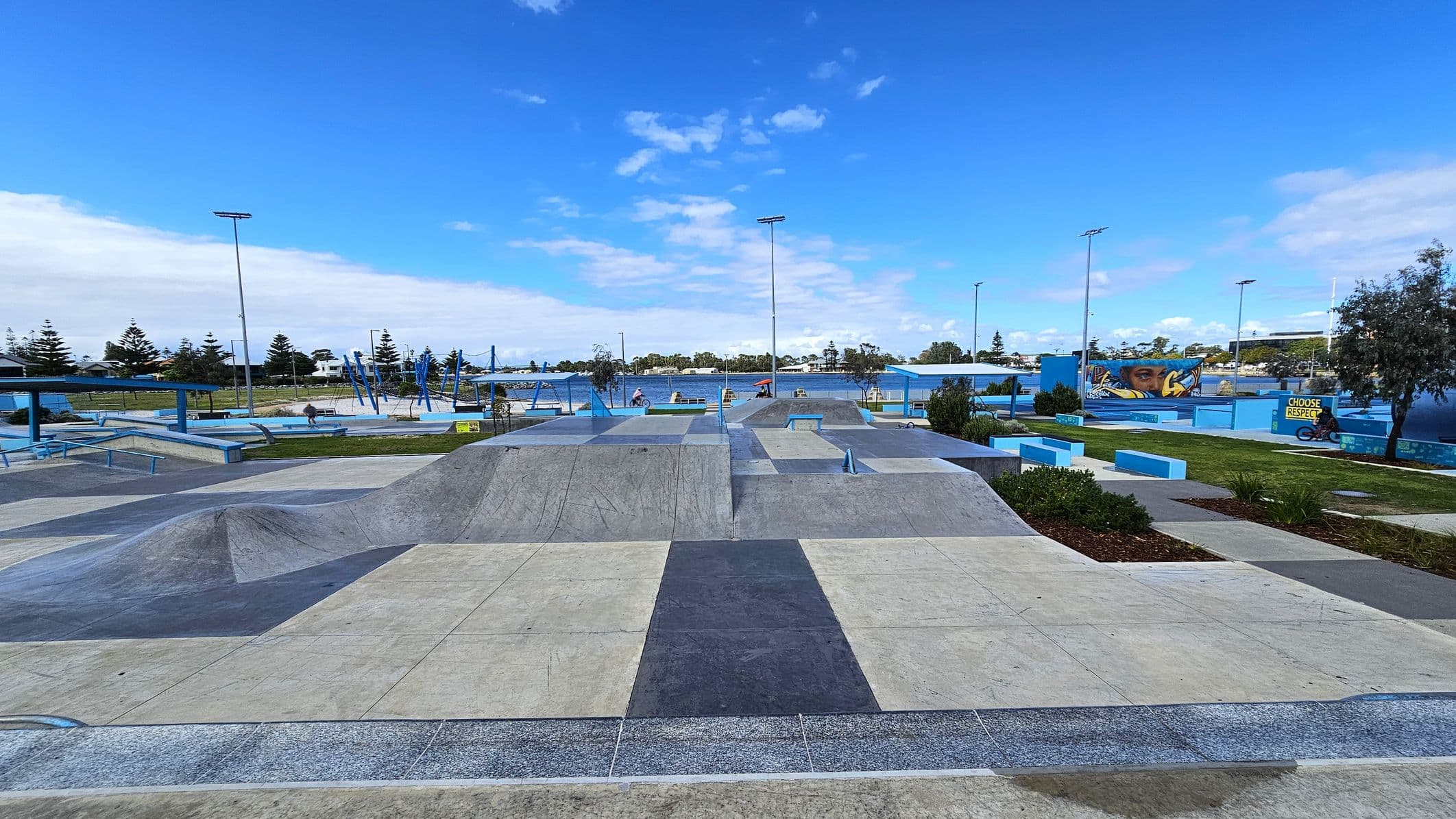 A skate park with various ramps and rails in the foreground, featuring a mix of concrete and blue-painted surfaces. In the background, there are trees and a mural with a face on a wall. To the right, a person on a bicycle is near a sign that reads "CHOOSE RESPECT". The area is open and spacious, with scattered greenery and a clear sky above.