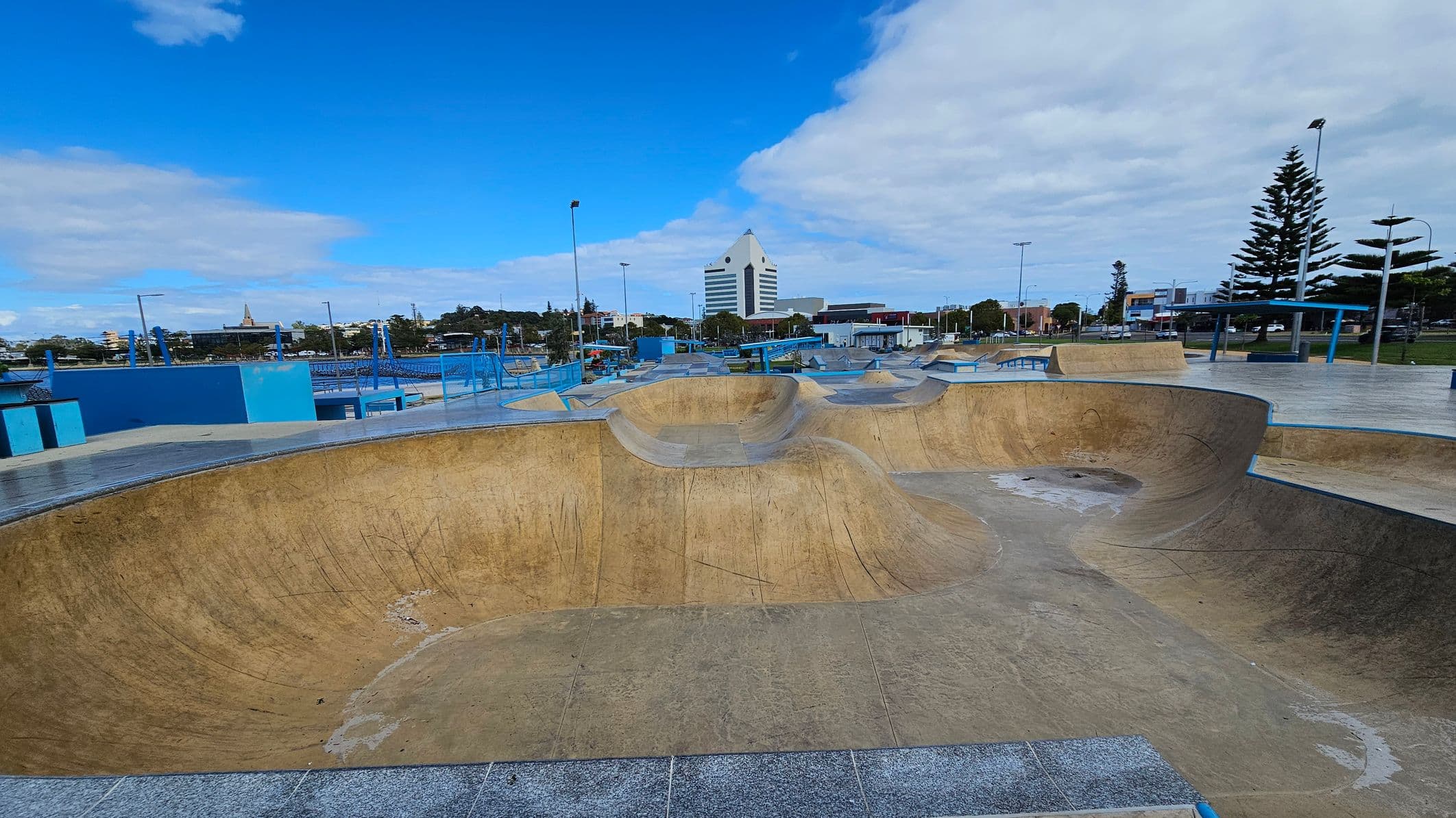 A skate park with large concrete bowls and ramps in the foreground. The surface is worn with visible marks. In the background, there is a distinctive white building with a triangular top. To the right, tall pine trees and street lamps line the area. Blue railings and benches are scattered throughout the park. The sky is partly cloudy, and the surrounding area includes various buildings and greenery.