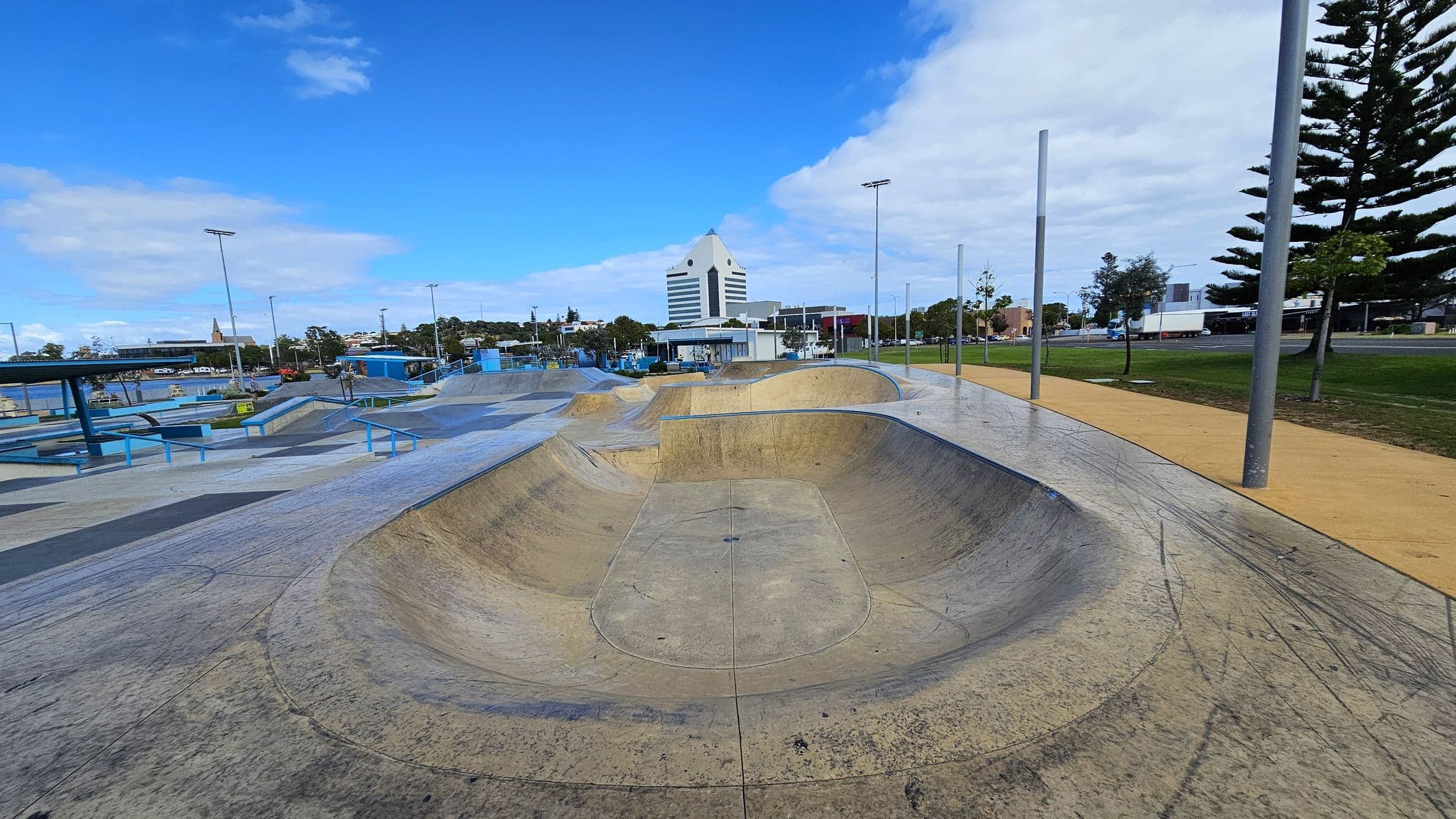 A skate park with a large concrete bowl in the foreground, featuring various ramps and rails. On the left, there are blue structures and benches. In the background, a distinctive white building with a triangular top is visible, surrounded by trees and other buildings. On the right, there is a grassy area with tall trees and a path lined with streetlights. The sky is mostly clear with some clouds.