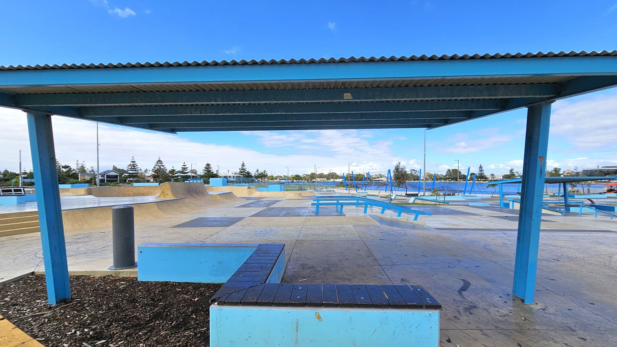 A skate park with various ramps and benches is visible. In the foreground, there's a blue metal shelter with a corrugated roof supported by blue pillars. The ground is a mix of concrete and wooden surfaces. In the background, there are trees and some buildings. Blue benches and additional structures are scattered throughout the park. The area is open and spacious, designed for skateboarding activities.