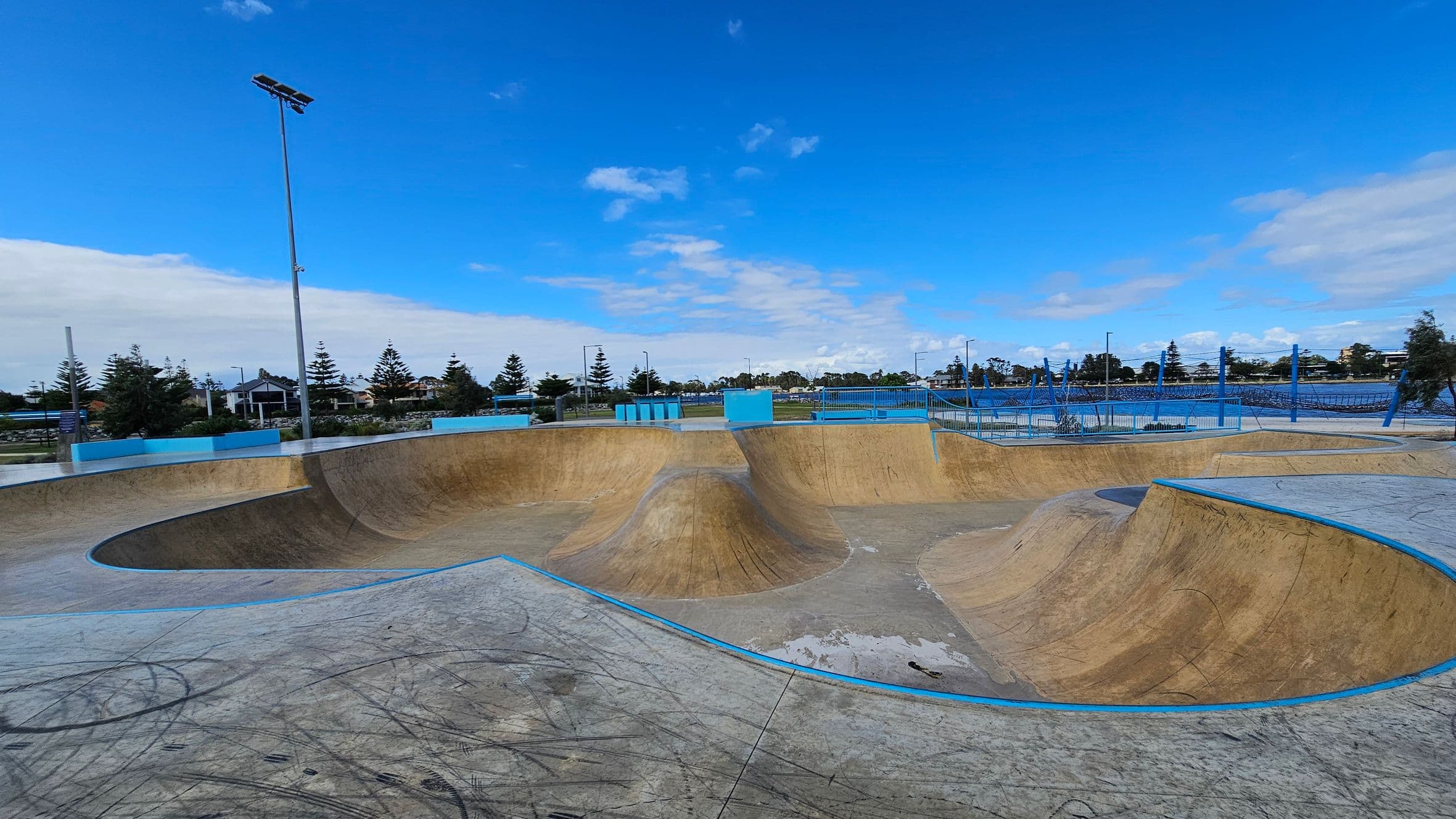 A skate park with multiple ramps and bowls in the foreground, featuring a smooth concrete surface with visible marks and scratches. The ramps are bordered with blue edges. In the background, there are tall floodlights and a fence enclosing the area. Beyond the fence, trees and buildings are visible, with a clear blue sky above.