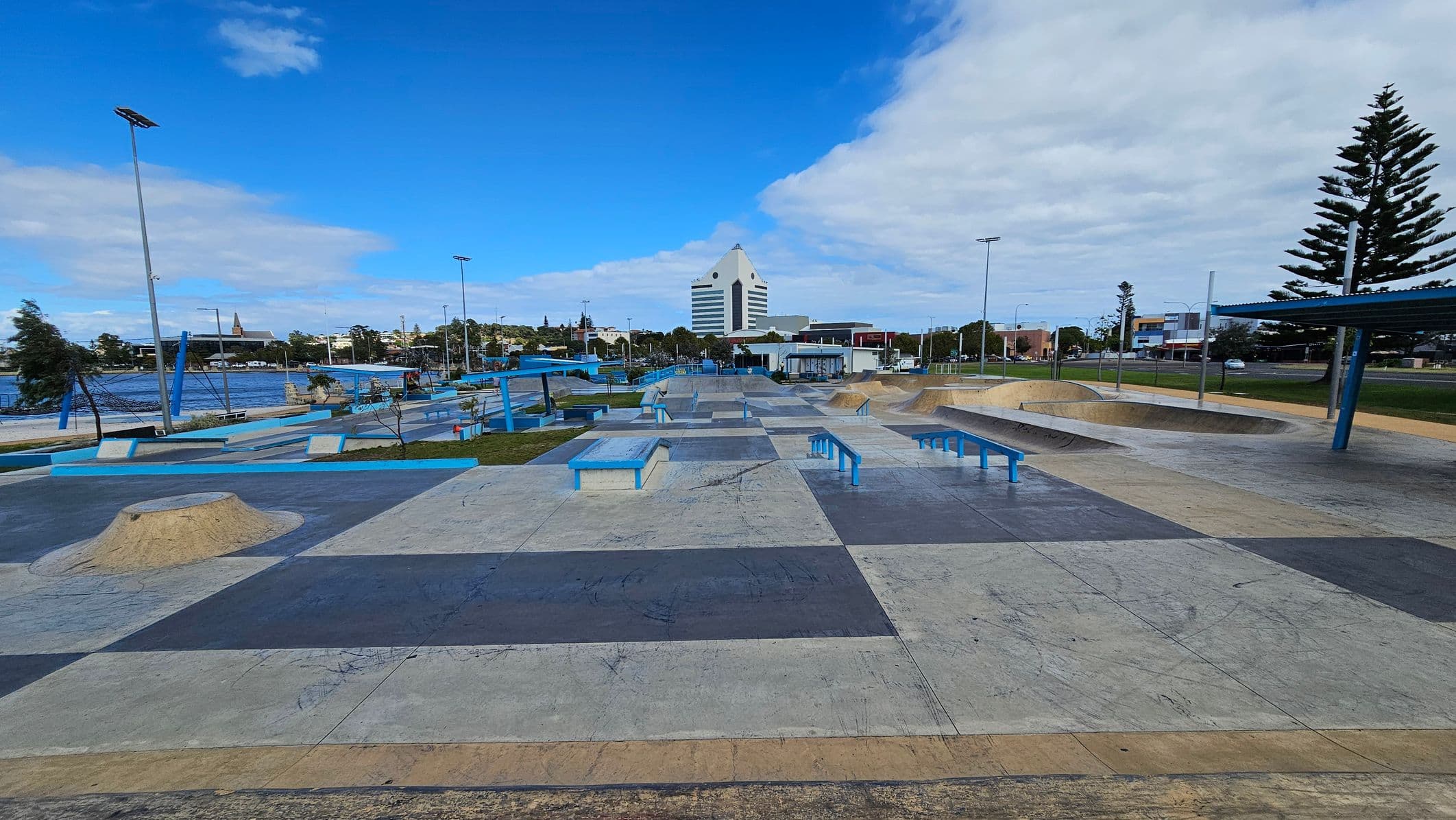 A skate park with various ramps, rails, and platforms in the foreground. The ground is a patchwork of different coloured concrete sections. In the background, there is a large, distinctive white building with a triangular top. To the left, there is a body of water with trees and a tall structure visible. On the right, there are more trees and a few buildings. The sky is partly cloudy, and there are tall streetlights throughout the area.