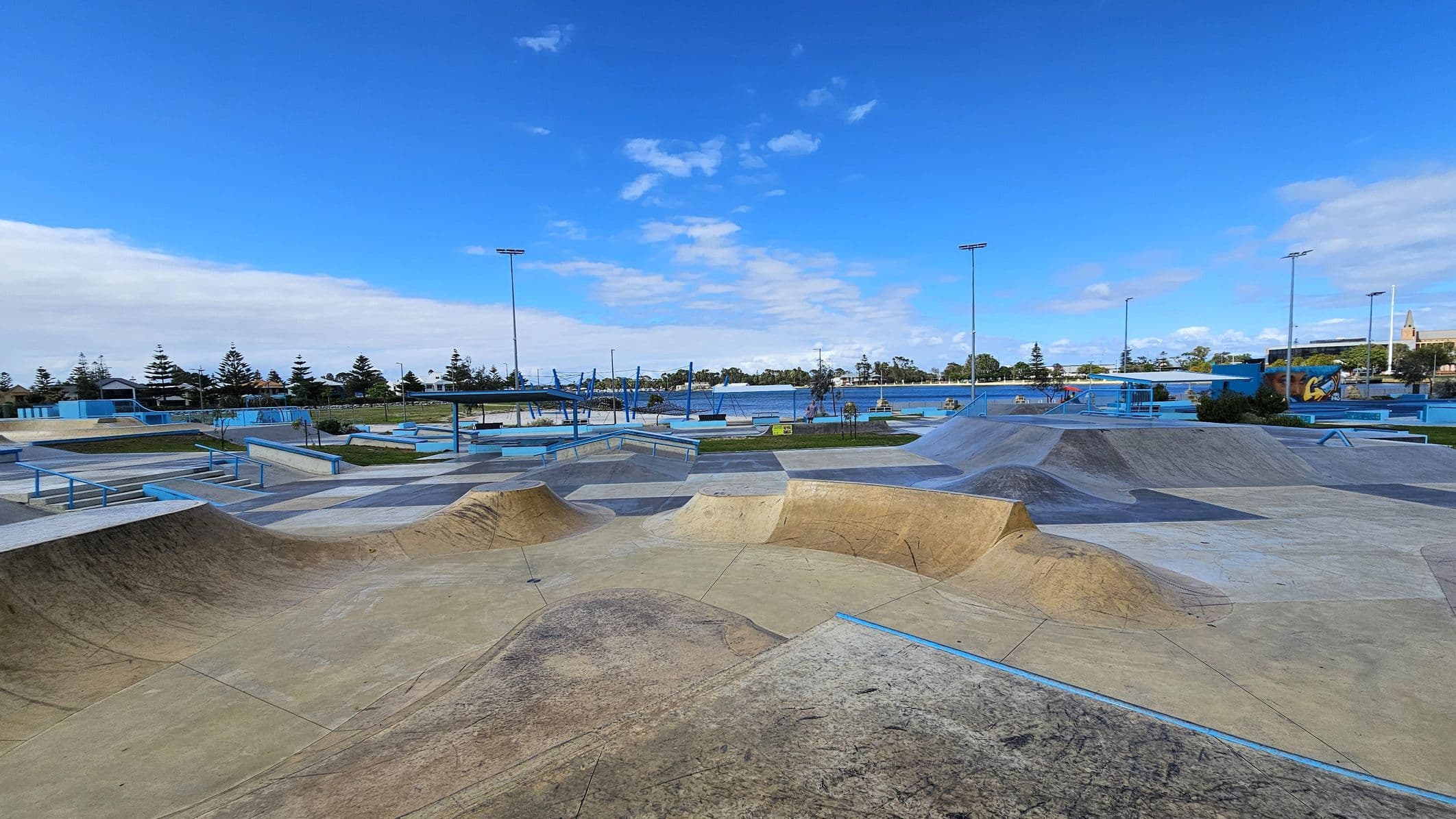 A skate park with various ramps and rails is in the foreground, featuring a mix of concrete and metal structures. The middle ground has more skate features and a few trees. In the background, there are tall light poles, a body of water, and a row of trees. A mural is visible on the right side. The sky is clear with some clouds.