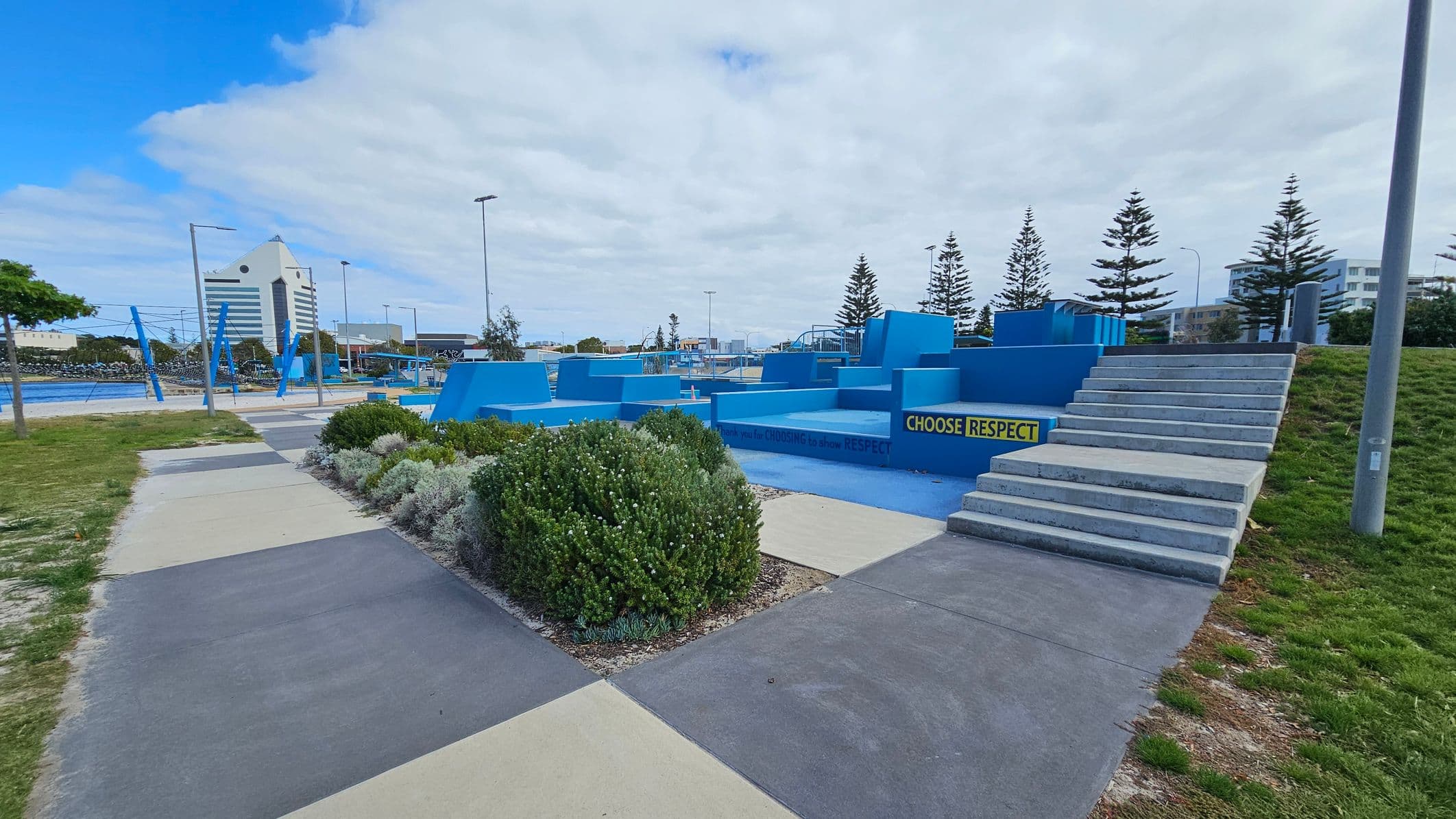 A skate park with blue ramps and structures is in the centre, featuring a sign that reads "CHOOSE RESPECT". On the right, there are concrete steps leading up to the park. In the foreground, there are patches of grass and a paved walkway with different coloured sections. In the background, a tall building with a triangular roof is visible, along with several pine trees and streetlights.