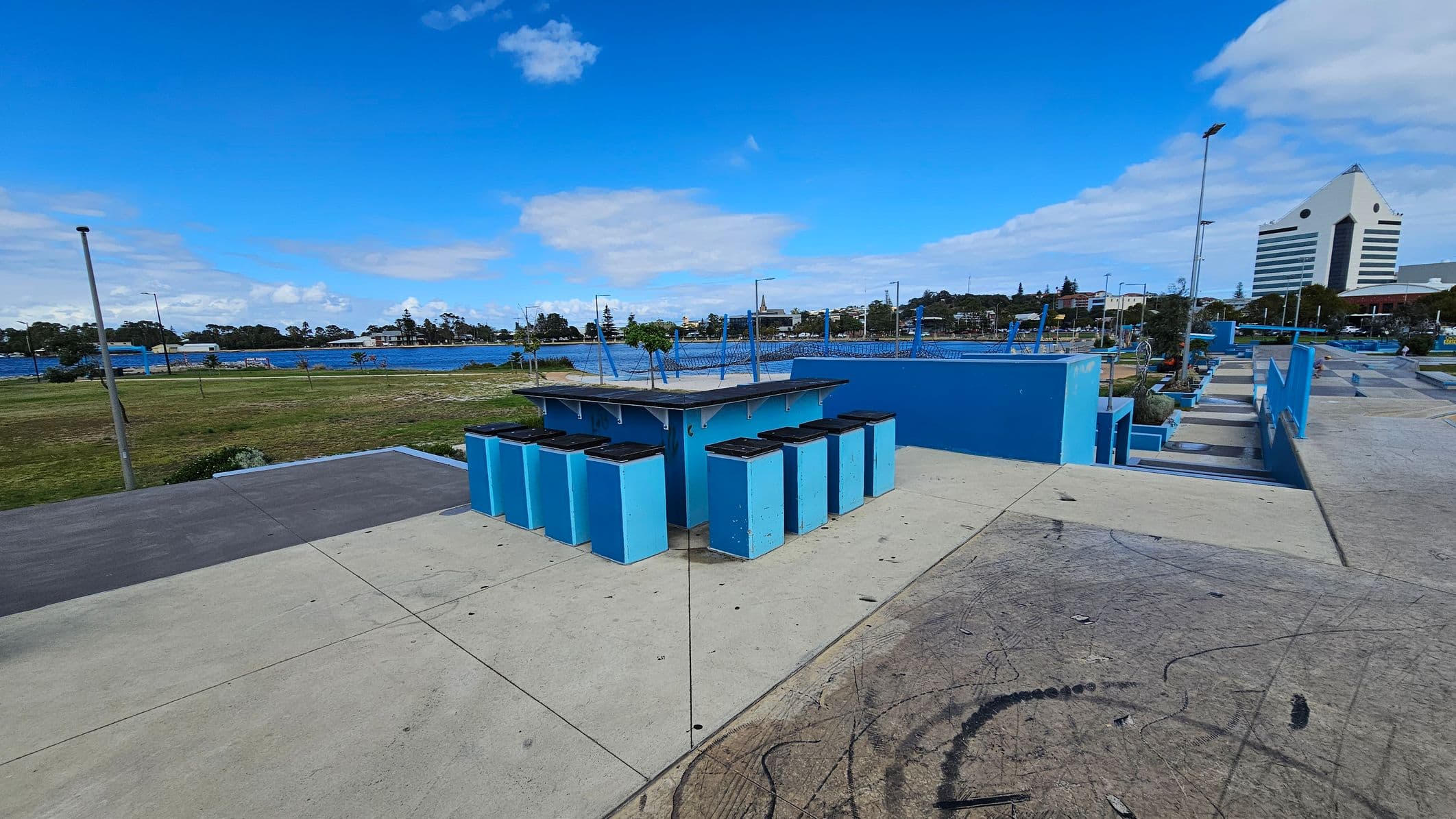 A concrete skate park with blue ramps and structures. In the foreground, there are several blue bins with black lids arranged in a semi-circle. The background features a grassy area leading to a body of water, with trees and buildings visible across the water. On the right, a distinctive white building with a triangular top stands out. The sky is clear with some clouds.