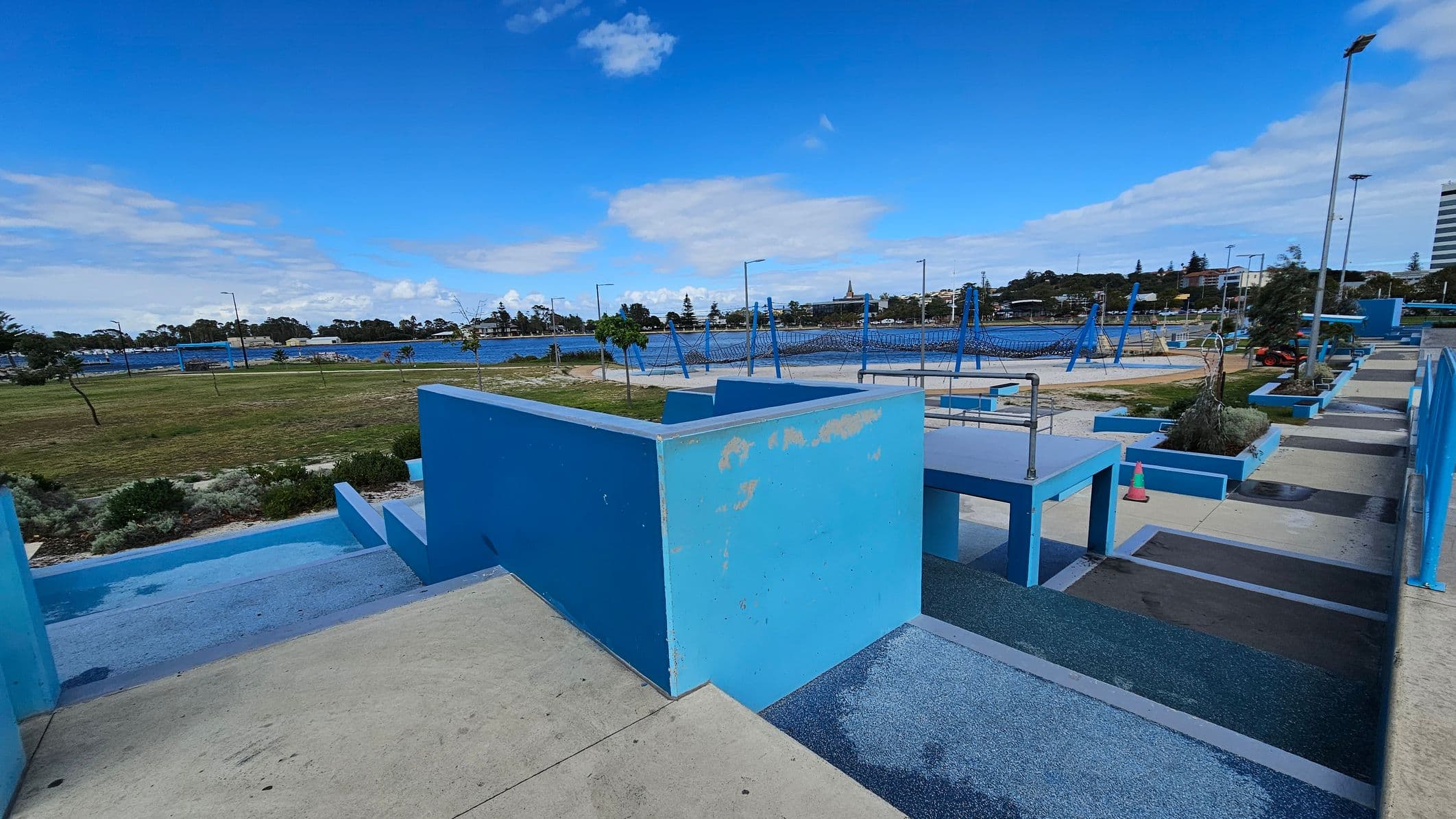 Concrete pathways and blue-painted structures are in the foreground, with a series of ramps and steps. In the middle, there are blue climbing frames and swings in a playground area. The background features a grassy field with scattered trees, a body of water, and a distant view of buildings and trees. The sky is mostly clear with some clouds.