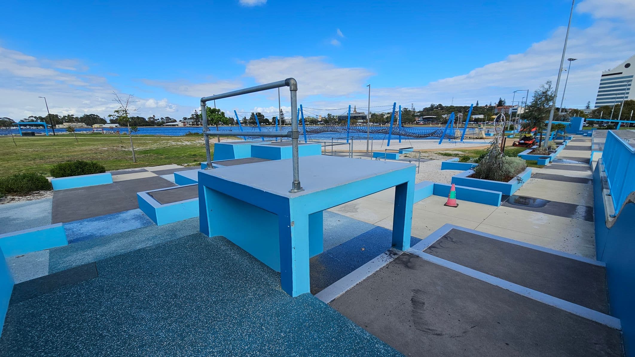 A parkour training area with various blue structures and platforms. In the foreground, a large blue platform with a metal bar on top is visible. To the right, there are more platforms and a pathway with a traffic cone. In the background, a grassy area and a body of water can be seen, along with trees and a few buildings. The ground is a mix of textured surfaces and concrete, and the area is open and spacious.