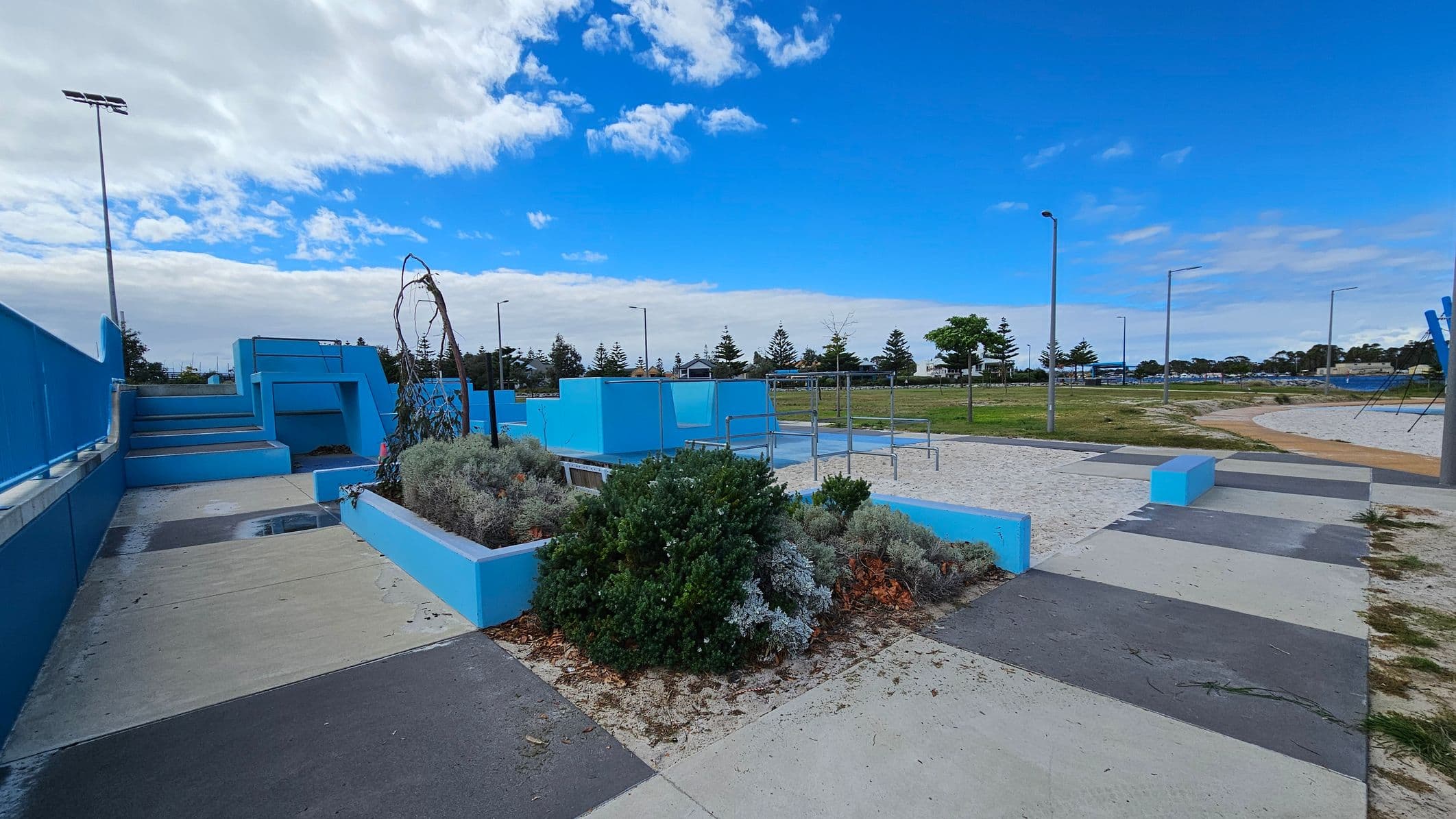 A park area with a blue and grey colour scheme. On the left, there are blue steps leading up to a platform with railings. In the centre, a raised garden bed with various shrubs and a small tree is surrounded by blue concrete borders. To the right, there is a sandy area with metal bars, possibly for exercise or play. The ground is a mix of concrete and sand, and there are several streetlights and trees in the background.