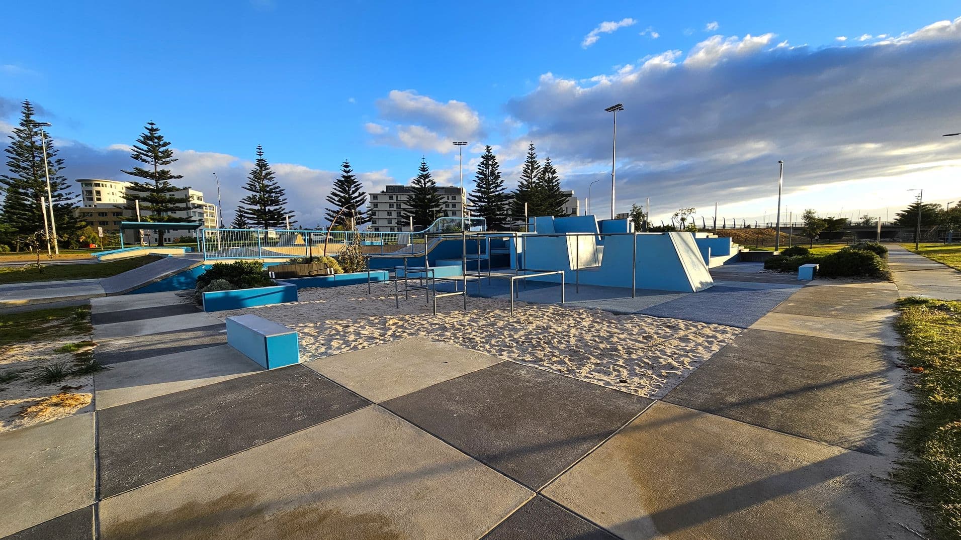 A skate park with various ramps and structures painted in blue and grey. The ground is a mix of concrete and sand. In the background, there are tall pine trees and several multi-storey buildings. The area is well-lit with tall street lamps, and there are pathways and grassy areas surrounding the park.
