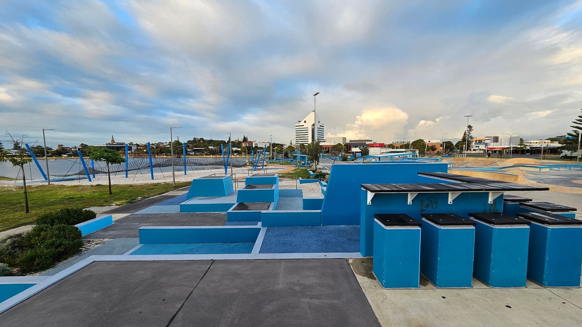 A skate park with blue ramps and structures is in the foreground, alongside a row of blue bins with black lids. In the background, there's a large building with a distinctive triangular roof. To the left, a sandy area with a netted climbing structure is visible, bordered by grass and small trees. The area is surrounded by various buildings and streetlights. The ground is a mix of concrete and blue surfaces.