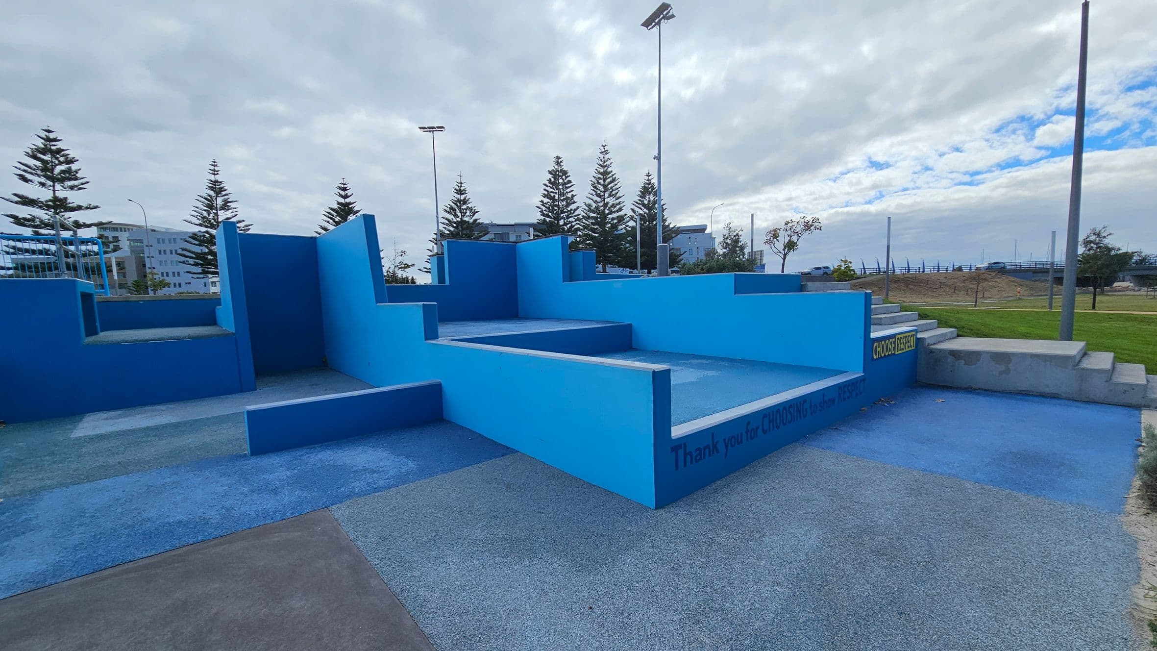 A geometric outdoor structure with various shades of blue, featuring multiple levels and steps. The ground is covered in textured, multicoloured surfaces. In the background, there are tall pine trees and a few buildings. A sign on the structure reads "Thank you for CHOOSING to show RESPECT." The area is open, with grassy patches and a few streetlights visible.