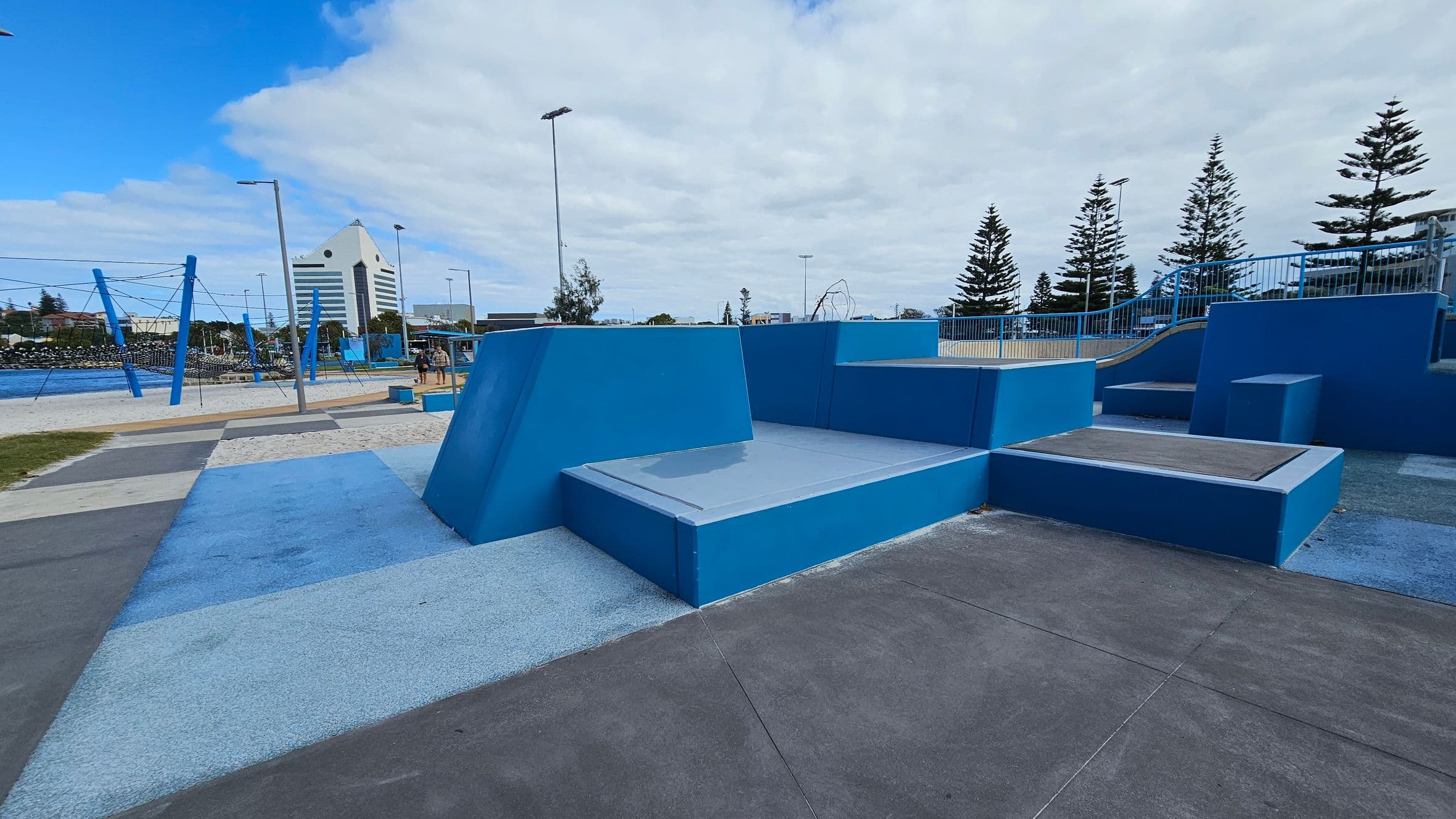 In the foreground, there are geometric structures in shades of blue and purple, resembling a skate park or playground. The ground is a mix of blue and purple surfaces. In the background, there are tall pine trees and a large, white, triangular-roofed building. To the left, there are blue poles and ropes, possibly part of a climbing structure, with a body of water visible beyond. The sky is overcast with patches of blue.
