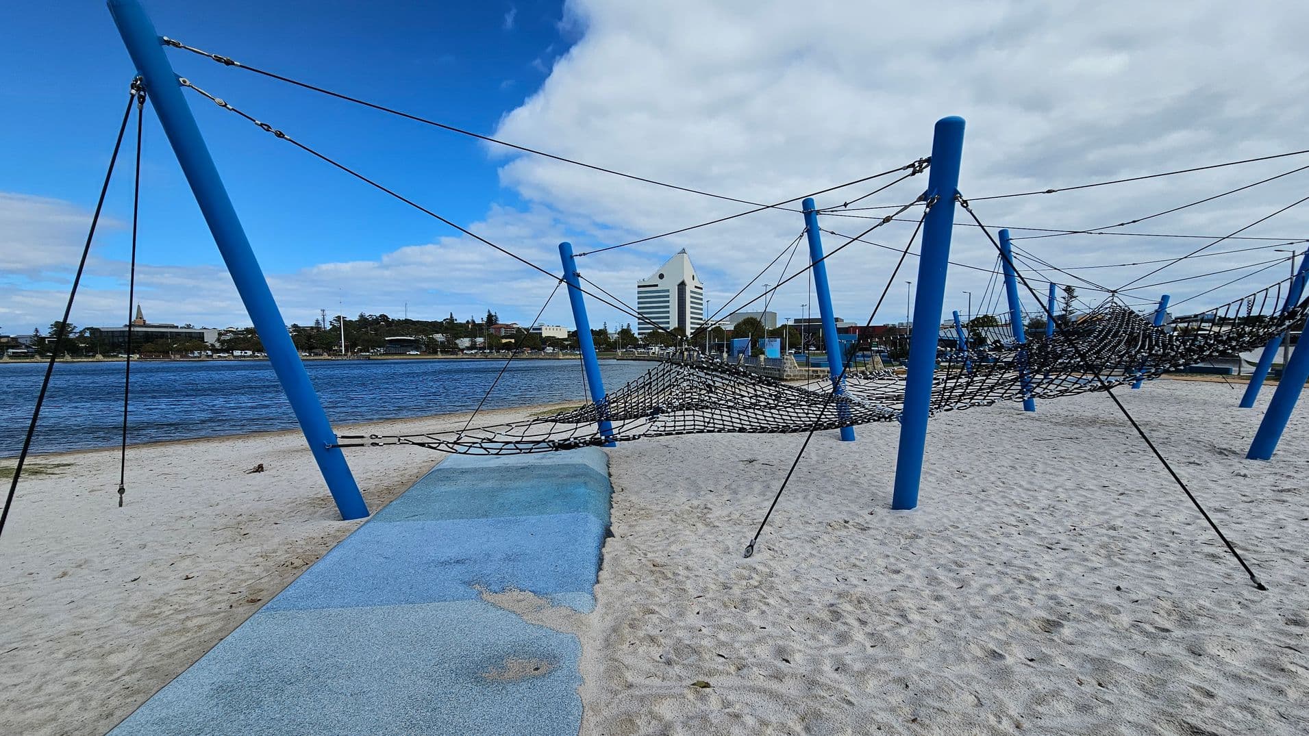A sandy playground with blue poles supporting a net climbing structure. The foreground has a blue rubber matting area. In the background, a body of water is visible with a cityscape beyond, including a distinctive white triangular-roofed building. The sky is partly cloudy.