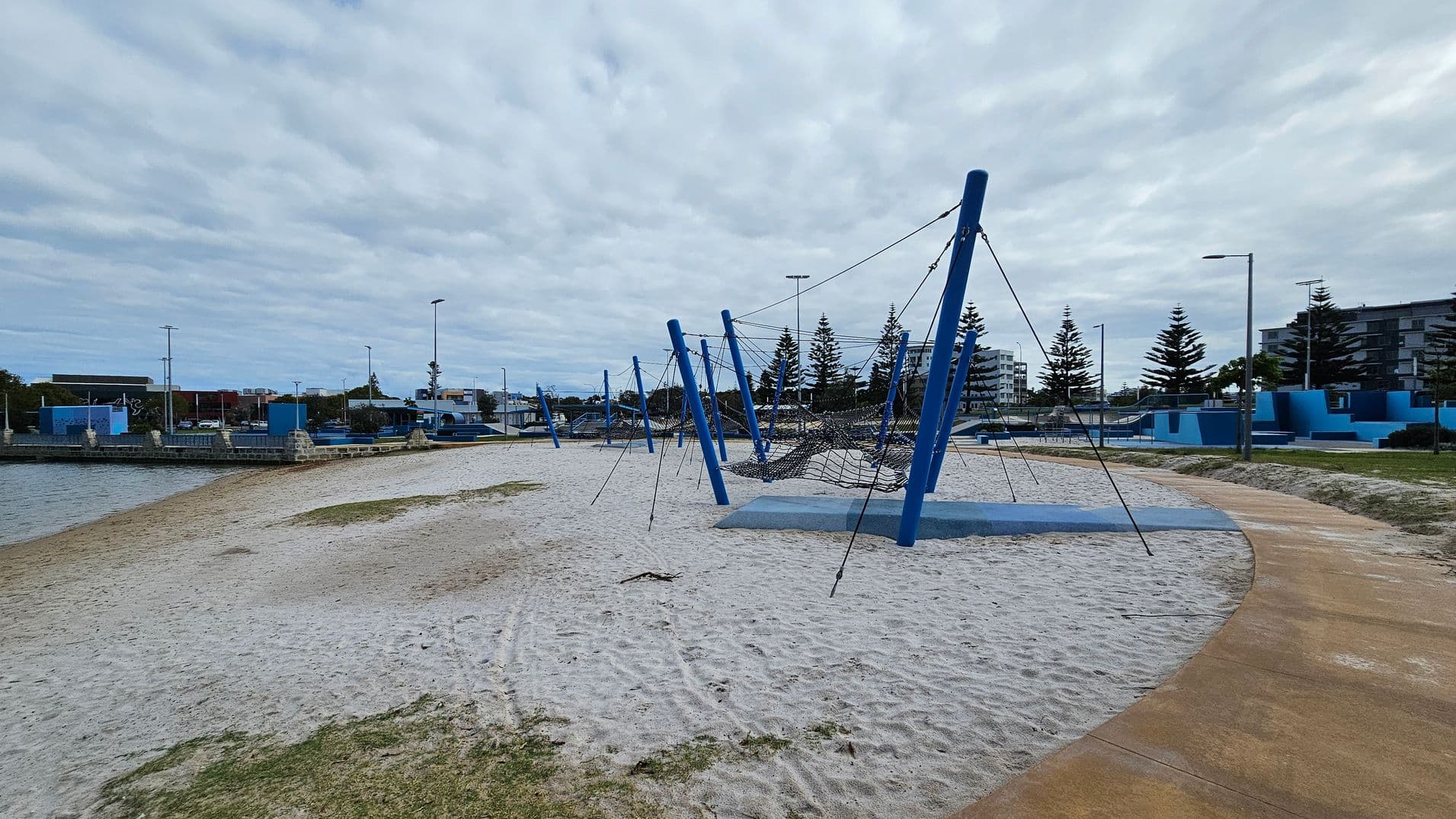 A sandy playground area with blue climbing structures and ropes is in the centre. On the left, there's a body of water bordered by a stone wall. In the background, there are several buildings and tall pine trees. A curved pathway runs along the right side, with grass patches scattered on the sand. Street lamps are visible throughout the area.