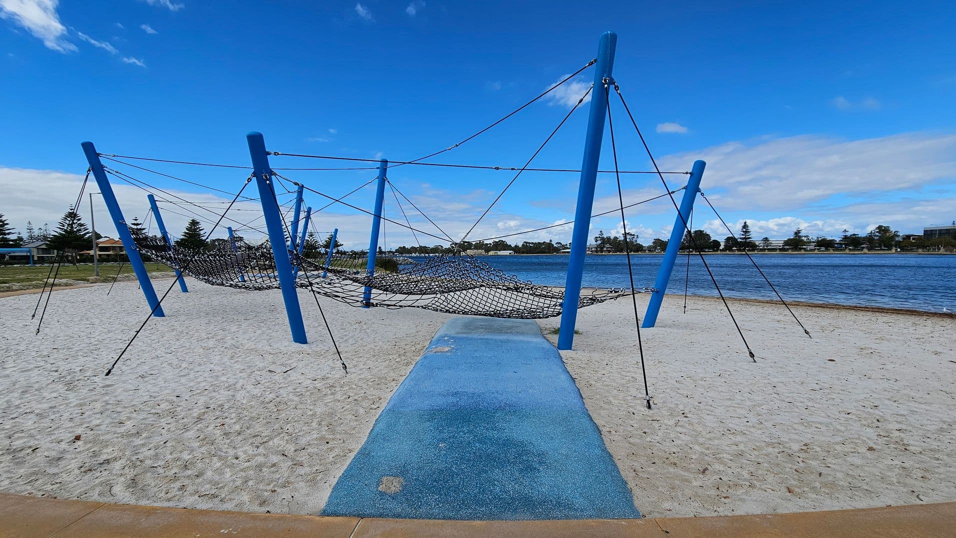A playground structure with blue poles and a netted climbing area is set on a sandy surface. A blue pathway leads to the structure. In the background, there is a body of water with a shoreline lined with trees and buildings. The sky is clear with a few clouds.
