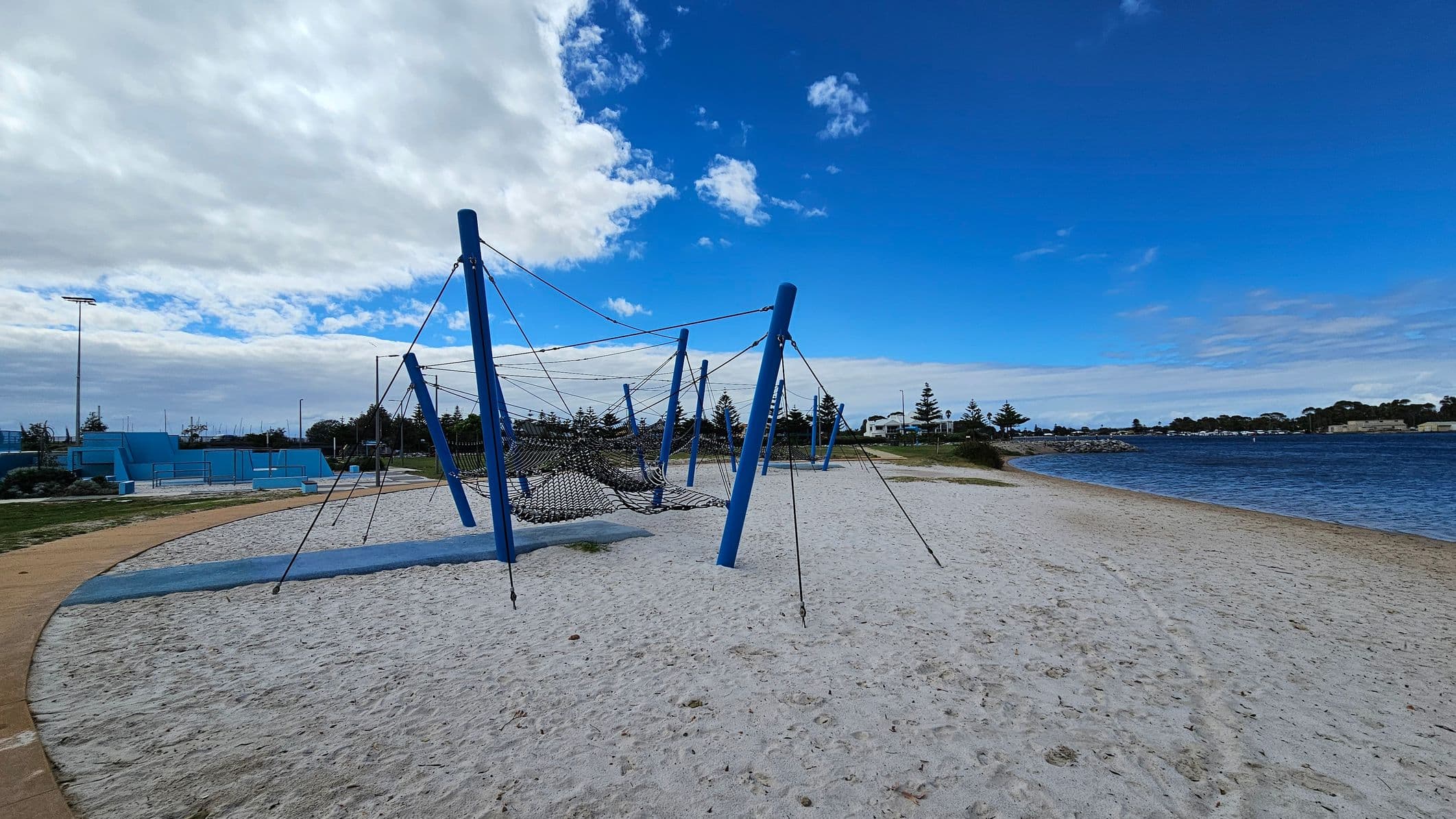 A sandy playground area with a large climbing structure made of blue poles and ropes is in the foreground. To the left, there are blue concrete structures and a pathway. In the background, there are trees and a body of water with a rocky shoreline. The sky is partly cloudy with patches of blue.