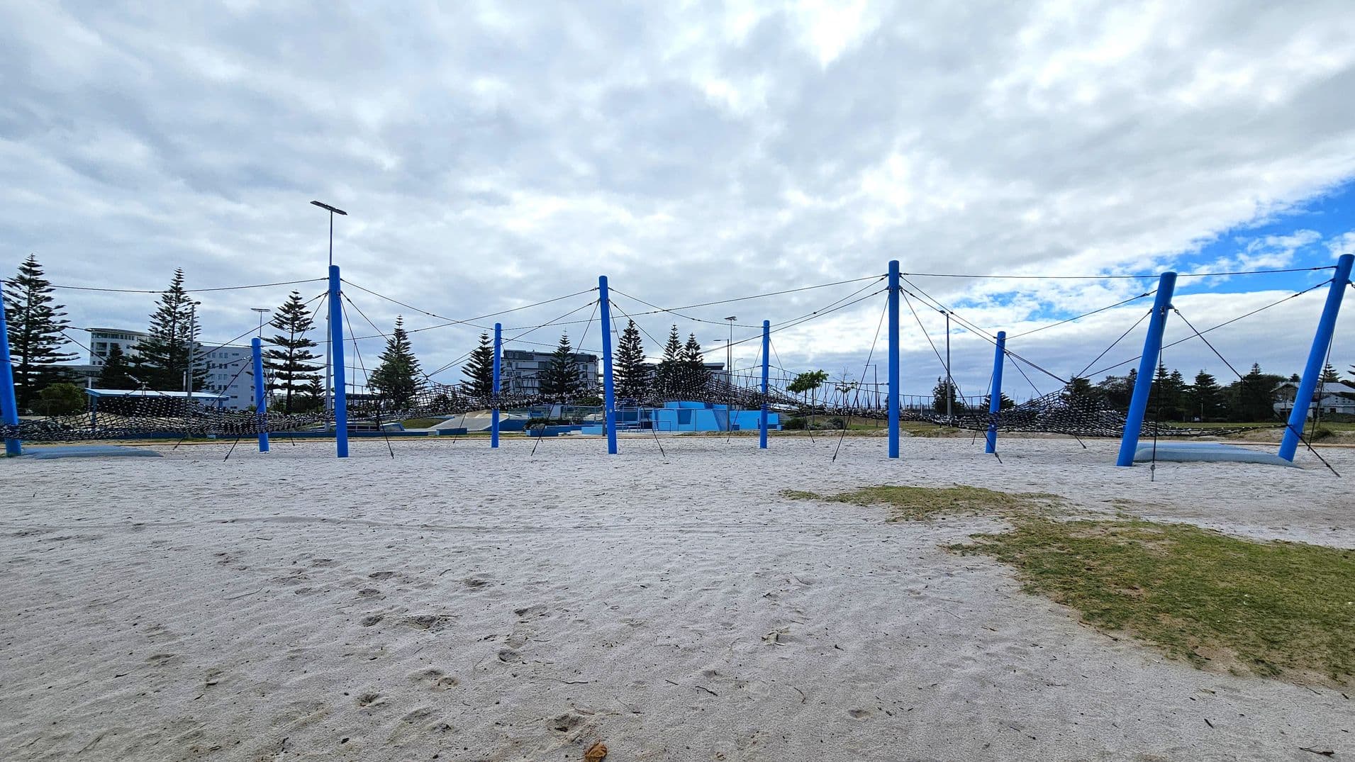 A sandy playground area with a large climbing structure made of blue poles and ropes. The structure is spread across the sand, with several poles connected by ropes forming a net-like design. In the background, there are trees and a few buildings visible. The ground has patches of grass mixed with the sand. The sky is overcast with some blue patches.