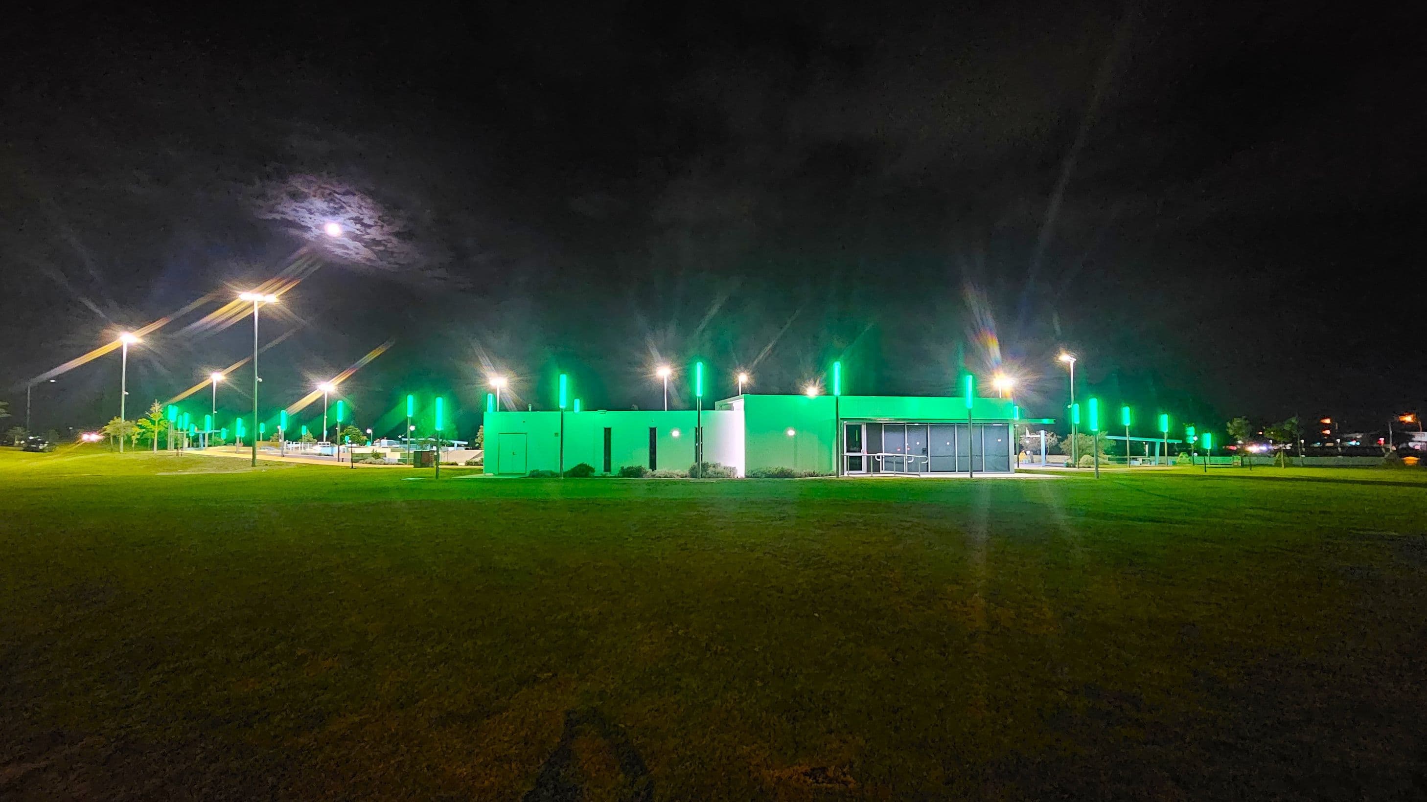 A modern building is centrally located, illuminated with green lights. To the left, a row of street lamps lines a pathway, casting a warm glow. The foreground features a grassy area, while the background is a dark night sky with a bright moon partially obscured by clouds. The right side of the building has large glass windows, and there are more green-lit poles extending into the distance.