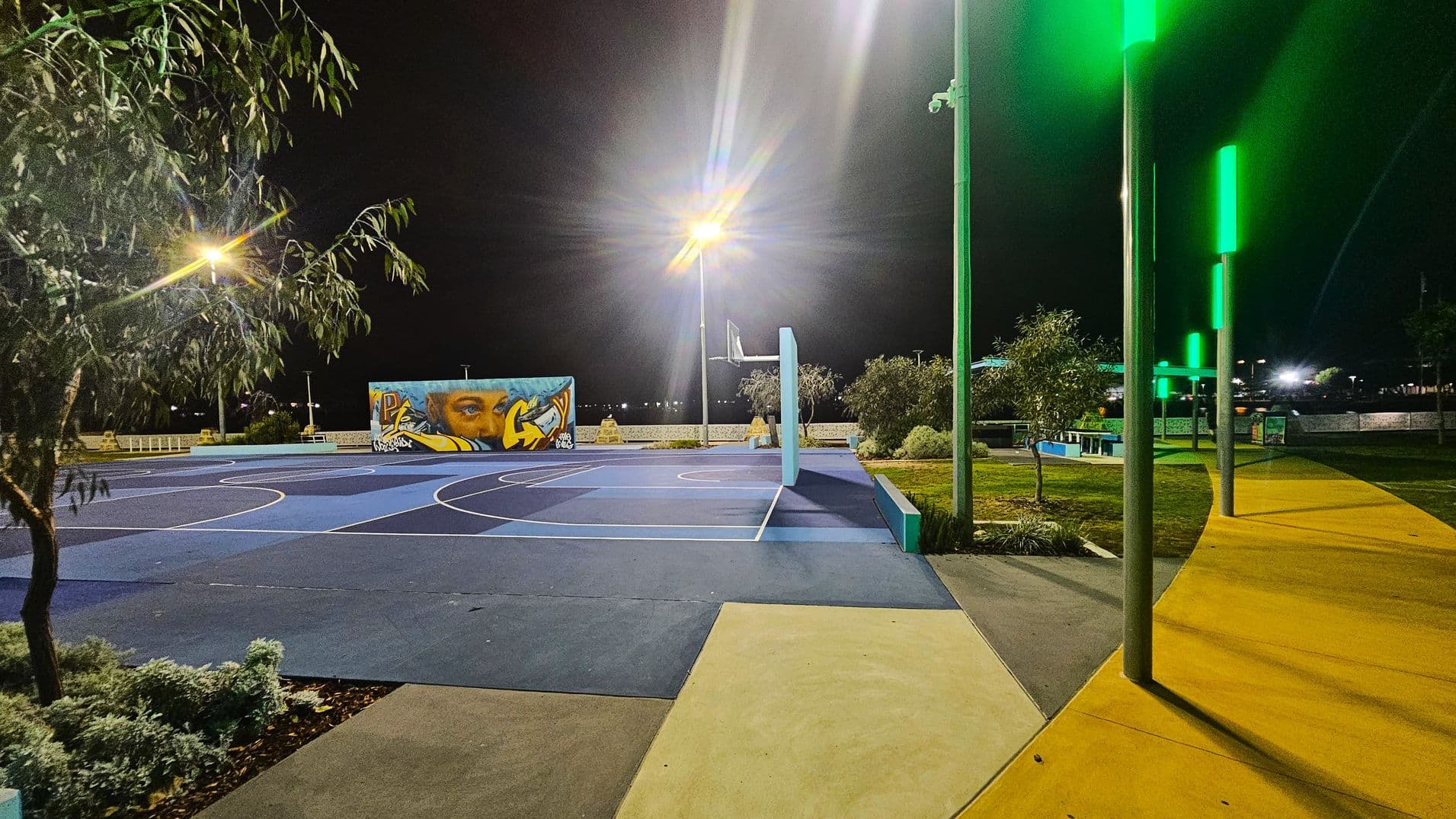 A night-time scene of an outdoor basketball court with a blue surface and white lines. On the left, a tree is visible under a bright streetlight. In the centre, a mural with a face and graffiti is painted on a wall. On the right, a path with yellow paving is lined with tall poles emitting green light. The area is surrounded by small bushes and trees, with additional lighting in the background.