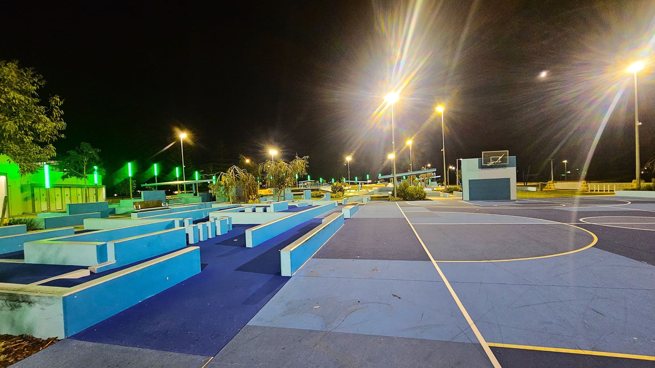 A night-time view of a sports area with a blue and purple basketball court on the right. The court features a hoop with a backboard. On the left, there are geometric blue structures, possibly for seating or parkour. Green lights illuminate the background, with trees and additional structures visible. Tall streetlights cast a warm glow over the scene. The ground is a mix of blue shades, and the sky is dark.
