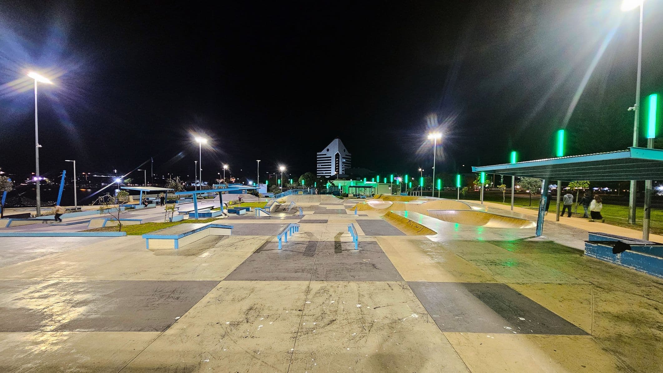 A well-lit skate park at night with various ramps and rails. The ground is a mix of concrete and coloured sections. On the right, there's a covered seating area with people standing nearby. Green lights line the path, and tall streetlights illuminate the area. In the background, a distinctive building with a triangular roof and striped facade is visible. The park is surrounded by trees and additional lighting.